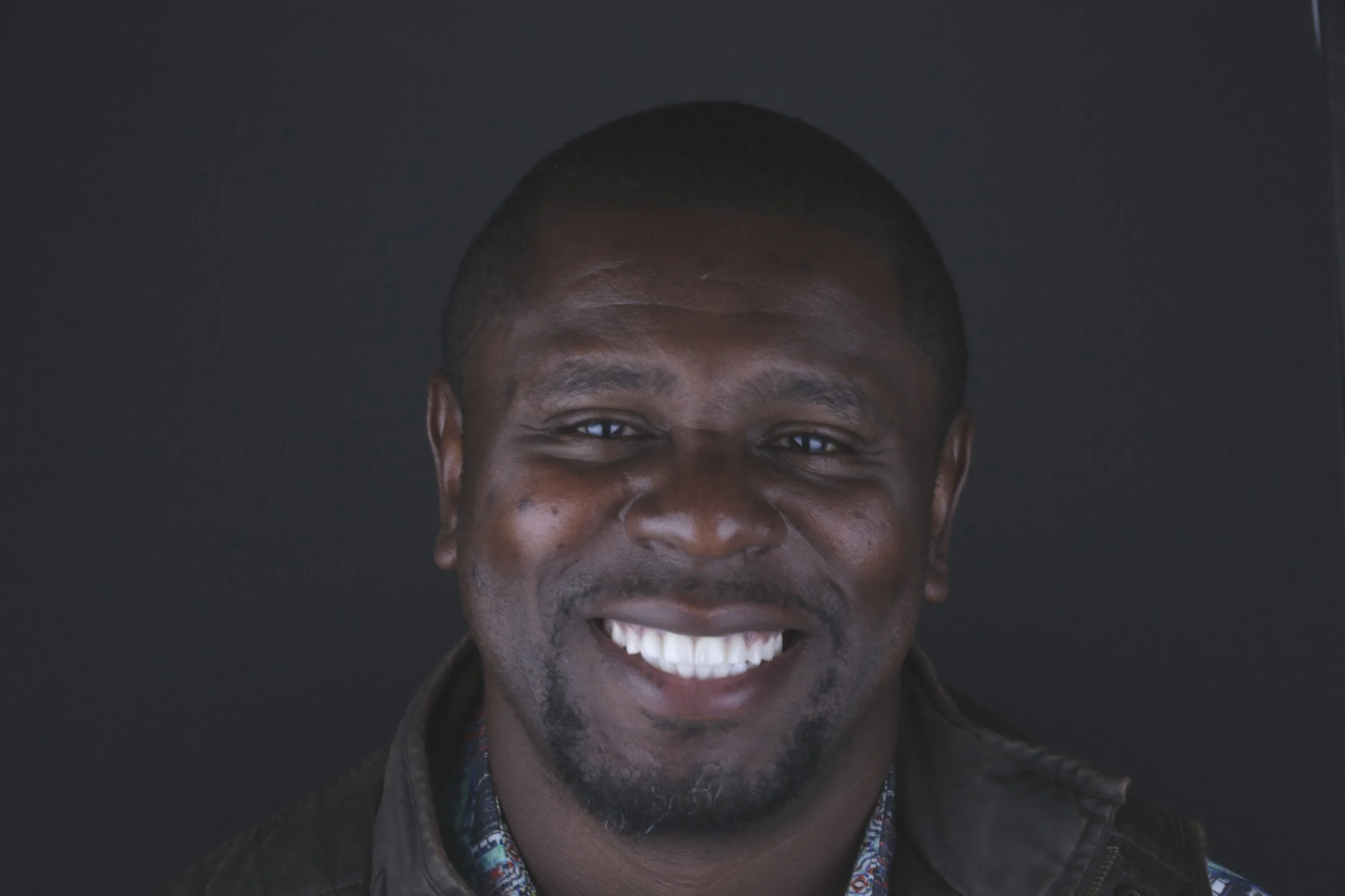 Close-up of a smiling man with short black hair, wearing a dark jacket and a patterned shirt, against a dark background.