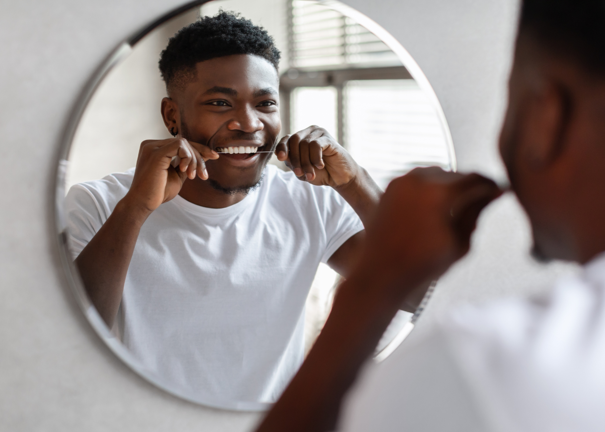 A young man is looking into a mirror while flossing his teeth and smiling.
