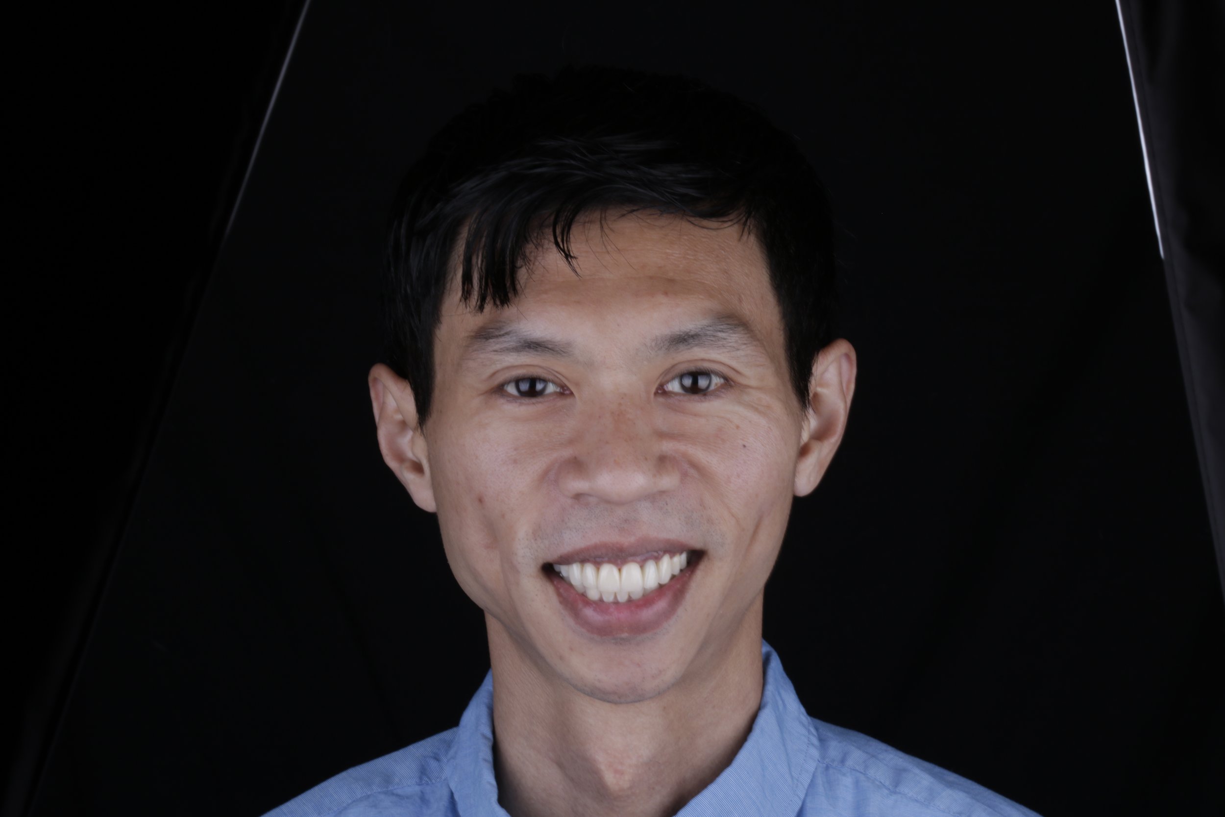 Close-up of a smiling man with short dark hair, wearing a blue collared shirt, against a dark background.