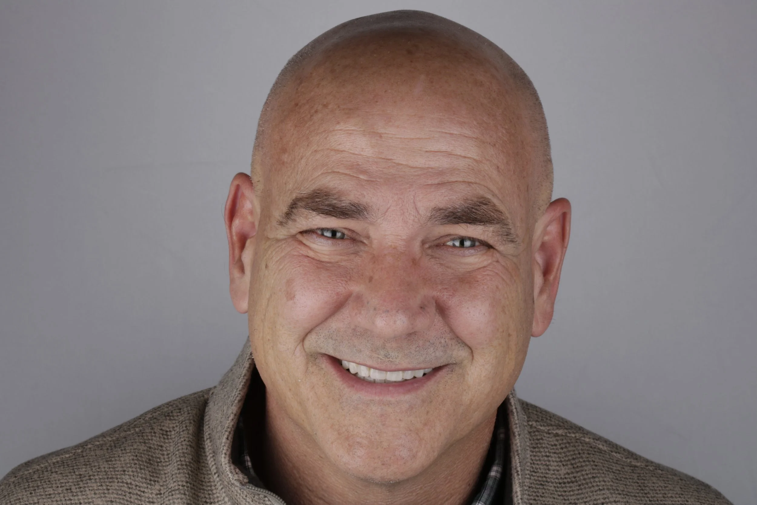 Close-up of a smiling man with light skin, bald head, gray eyes, and facial wrinkles, wearing a brown jacket and a checkered shirt, against a plain gray background.
