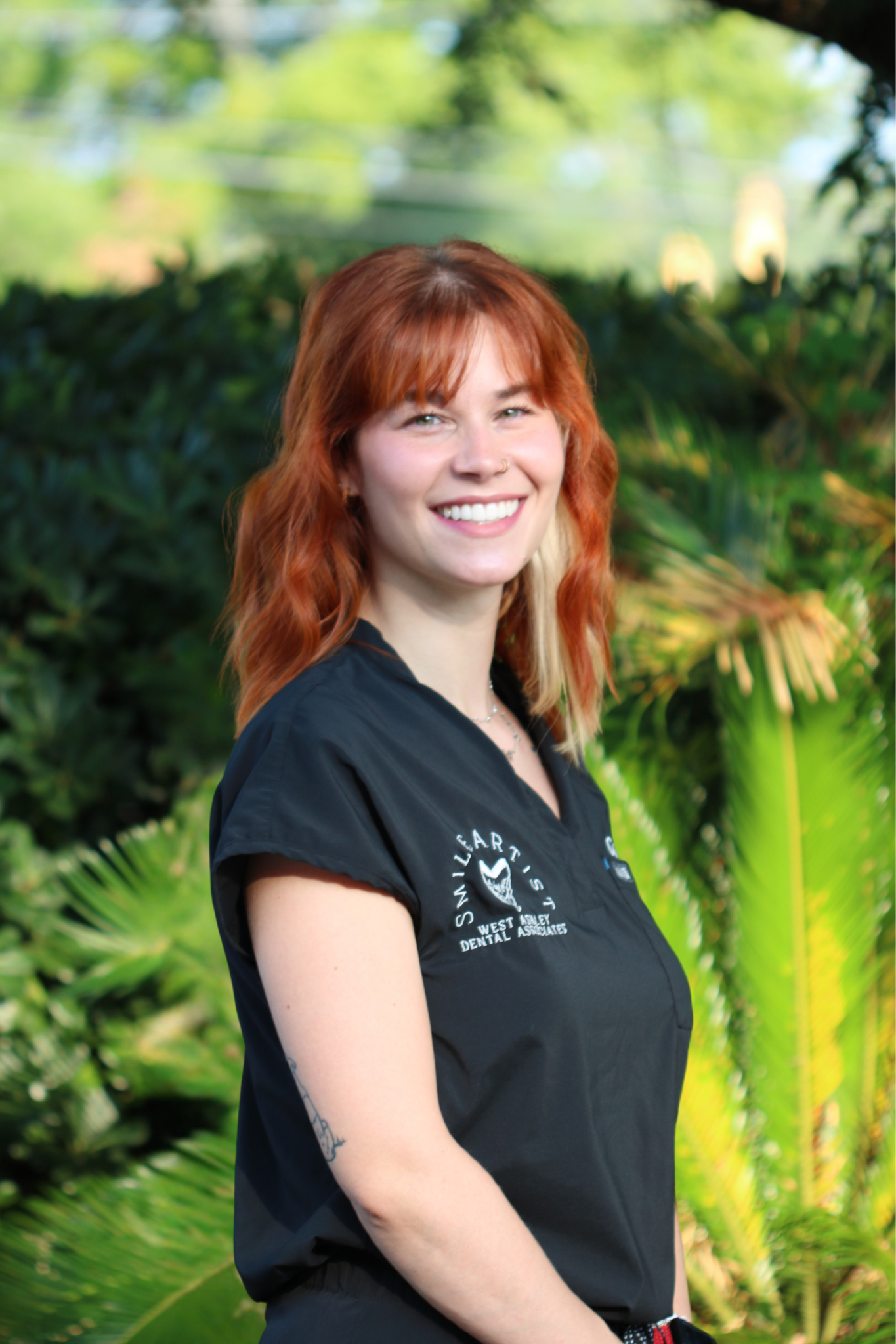 Close-up of a young woman with red hair, smiling, standing outdoors with green foliage in the background.