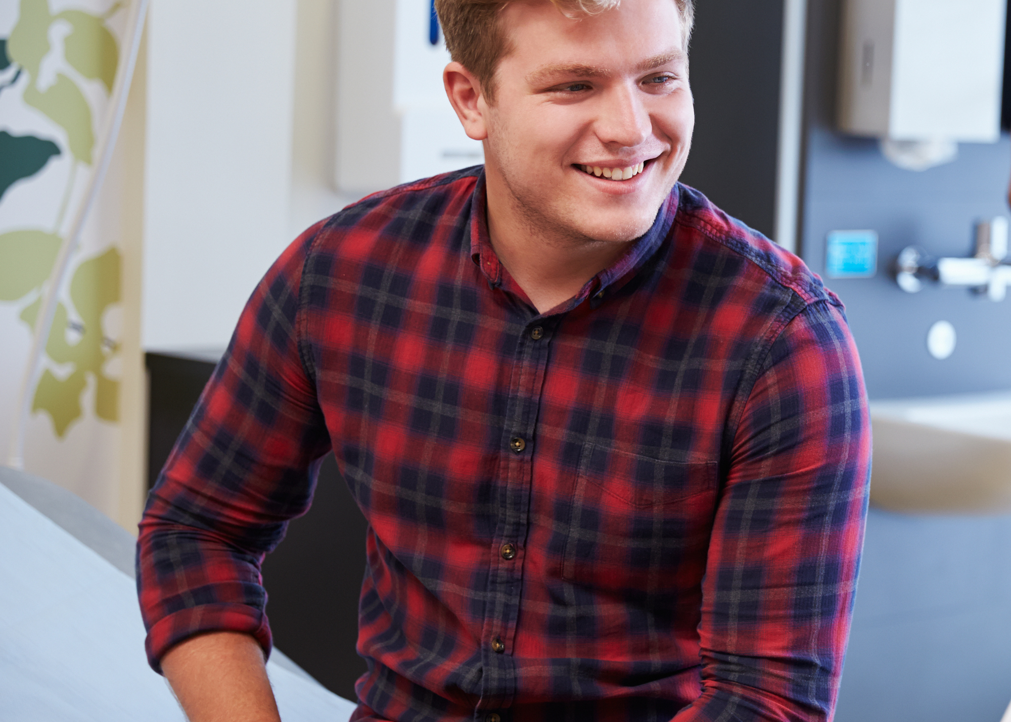 A young man with light skin and short reddish hair smiling, wearing a red and blue plaid shirt, in a modern medical or office setting.