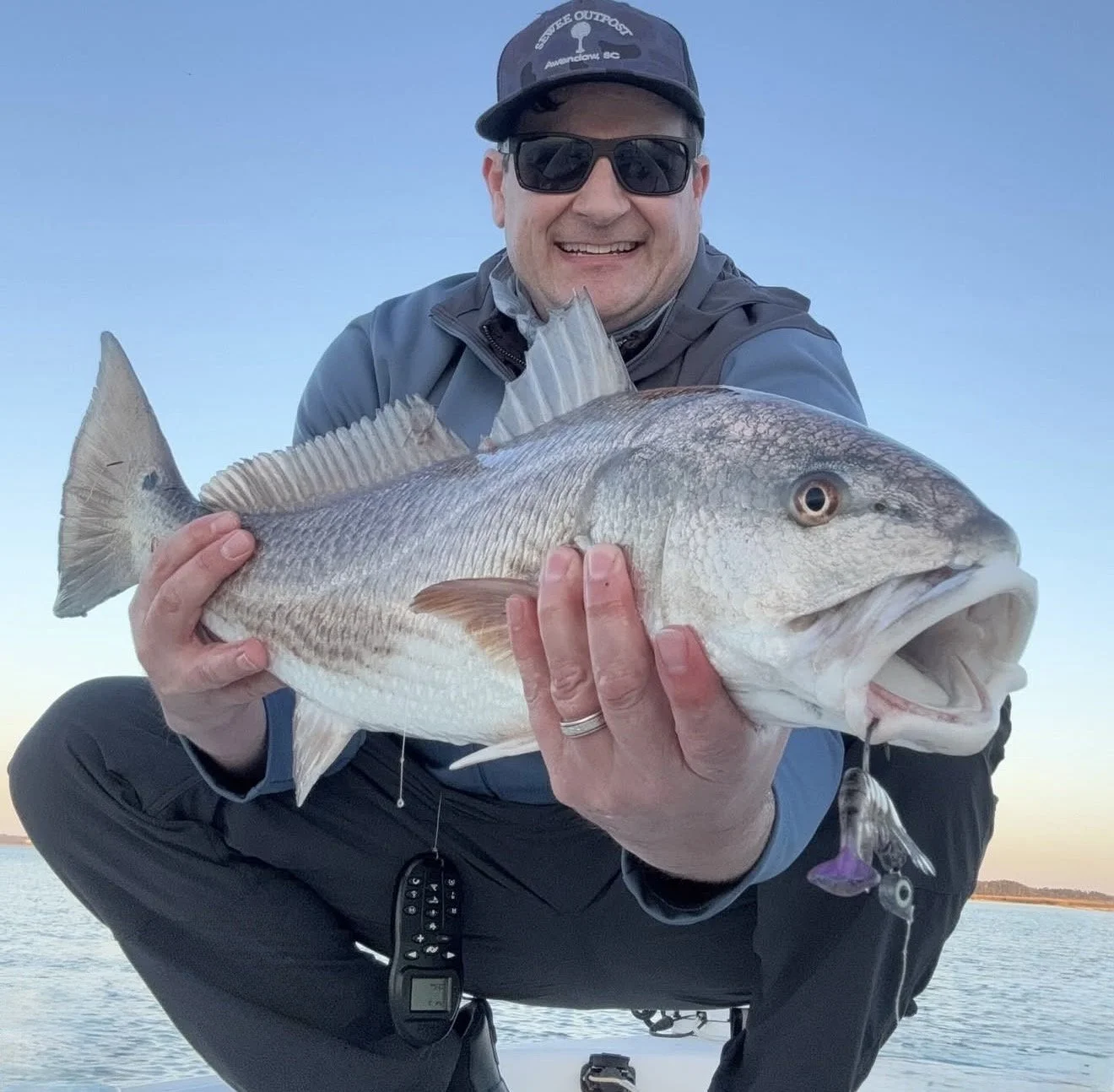 A smiling man wearing sunglasses and a cap, holding a large fish he caught, outdoors near water at dusk.