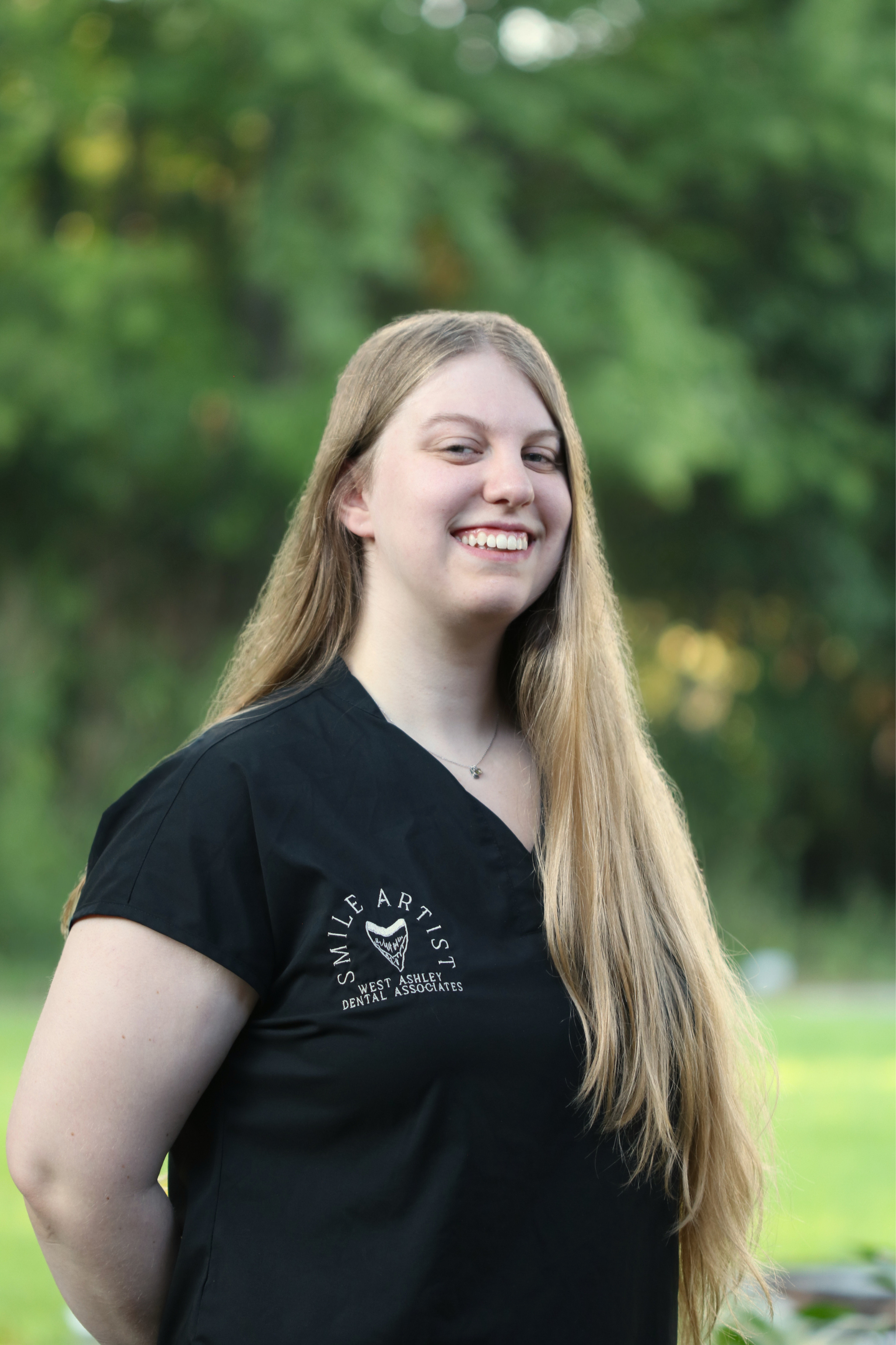 A young woman with long blonde hair smiling outdoors, wearing a black t-shirt with a dental practice logo and a necklace.