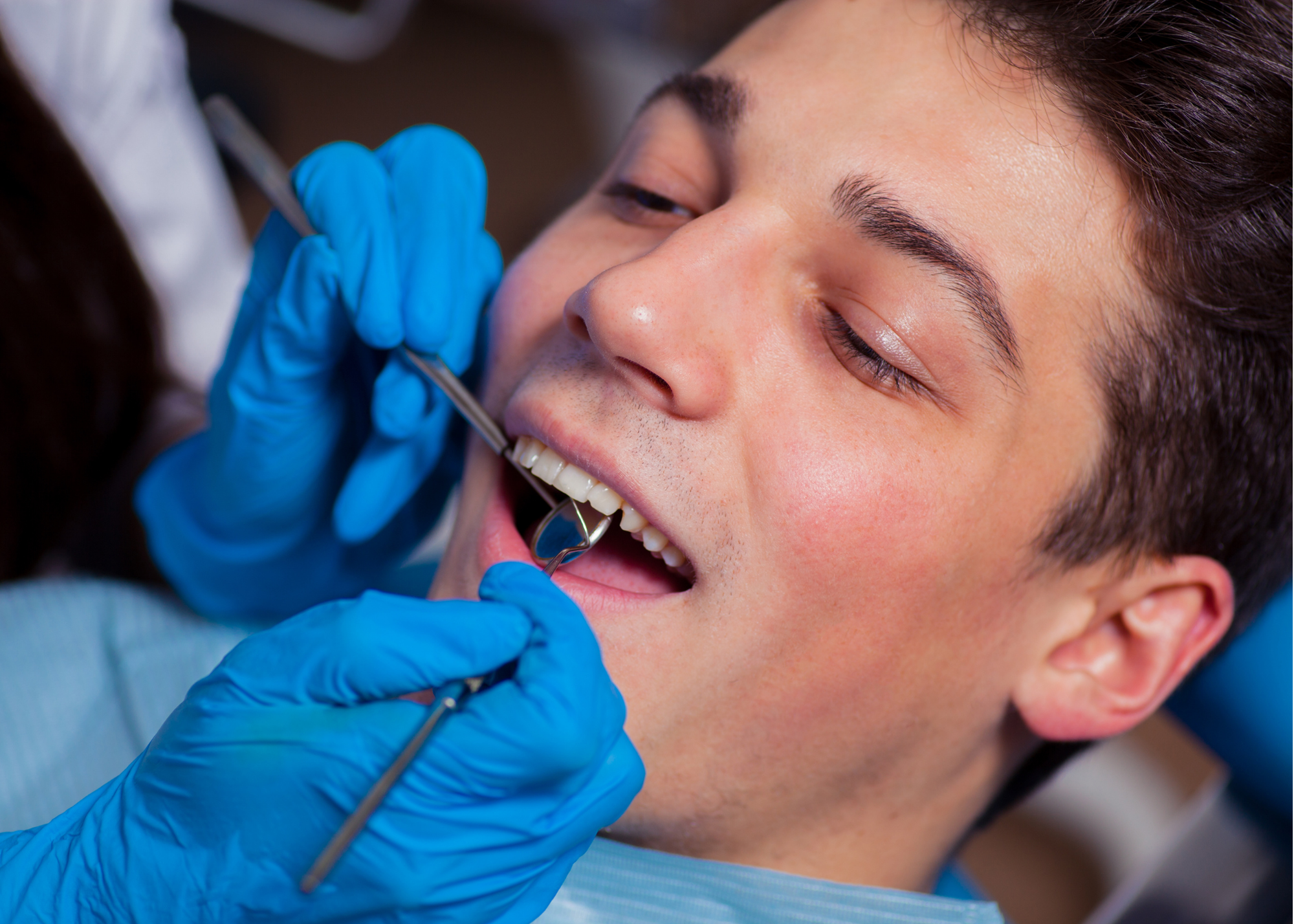 A young man receiving a dental examination from a dentist wearing blue gloves with a dental mirror in his mouth.