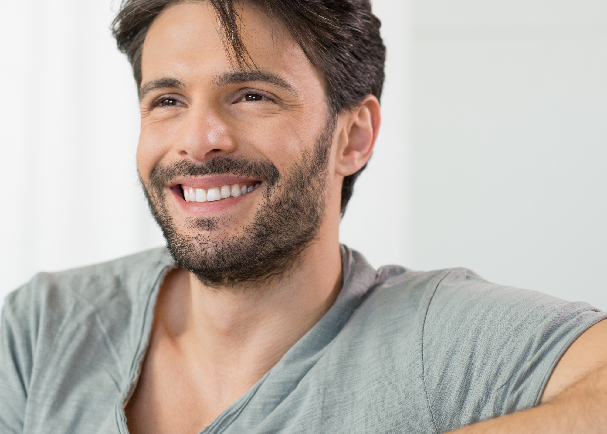 A smiling man with dark brown hair and a beard, wearing a gray shirt, taking a selfie in front of a white background.