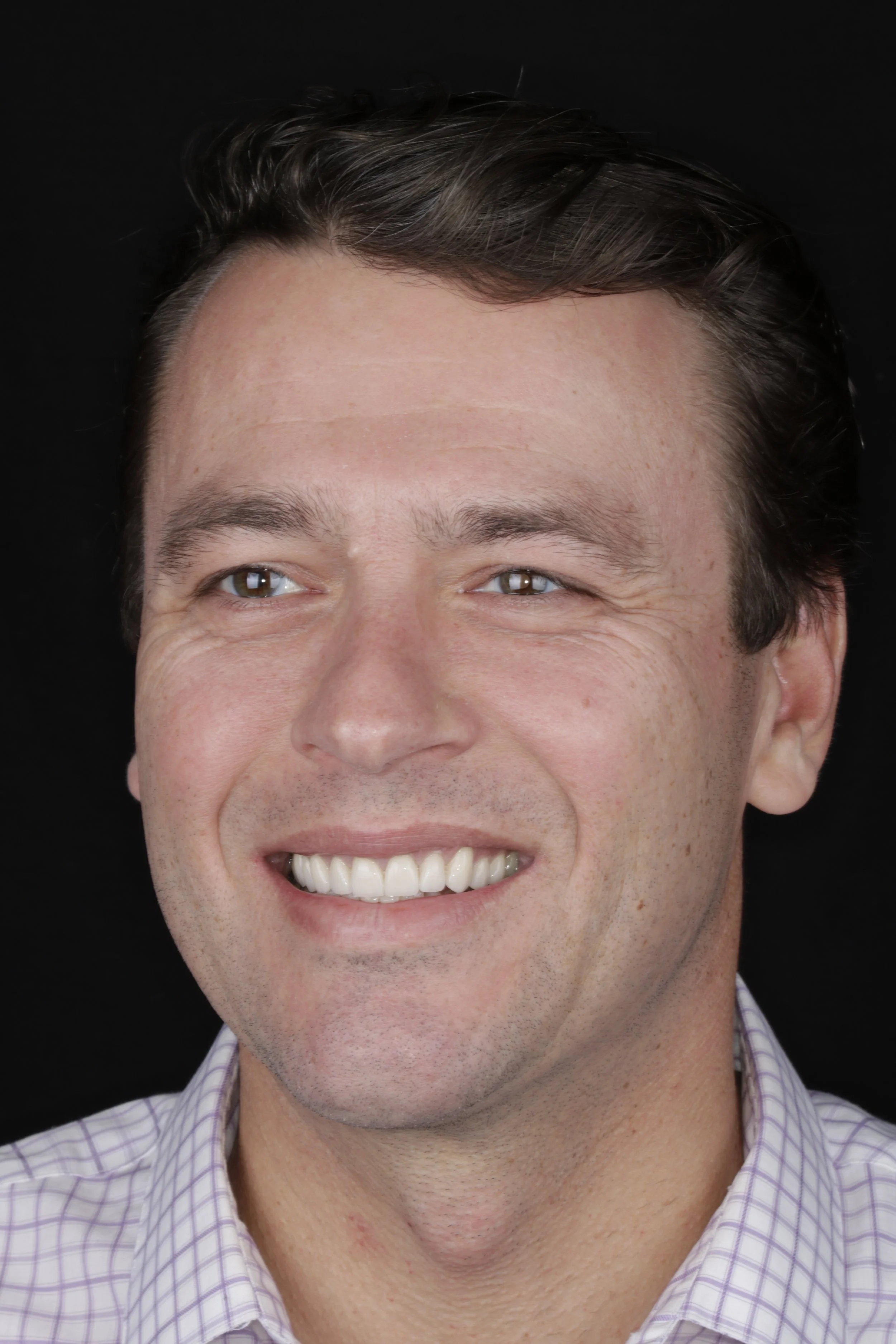 Close-up of a man smiling, showing his teeth, wearing a purple checkered shirt, with dark hair, against a black background.