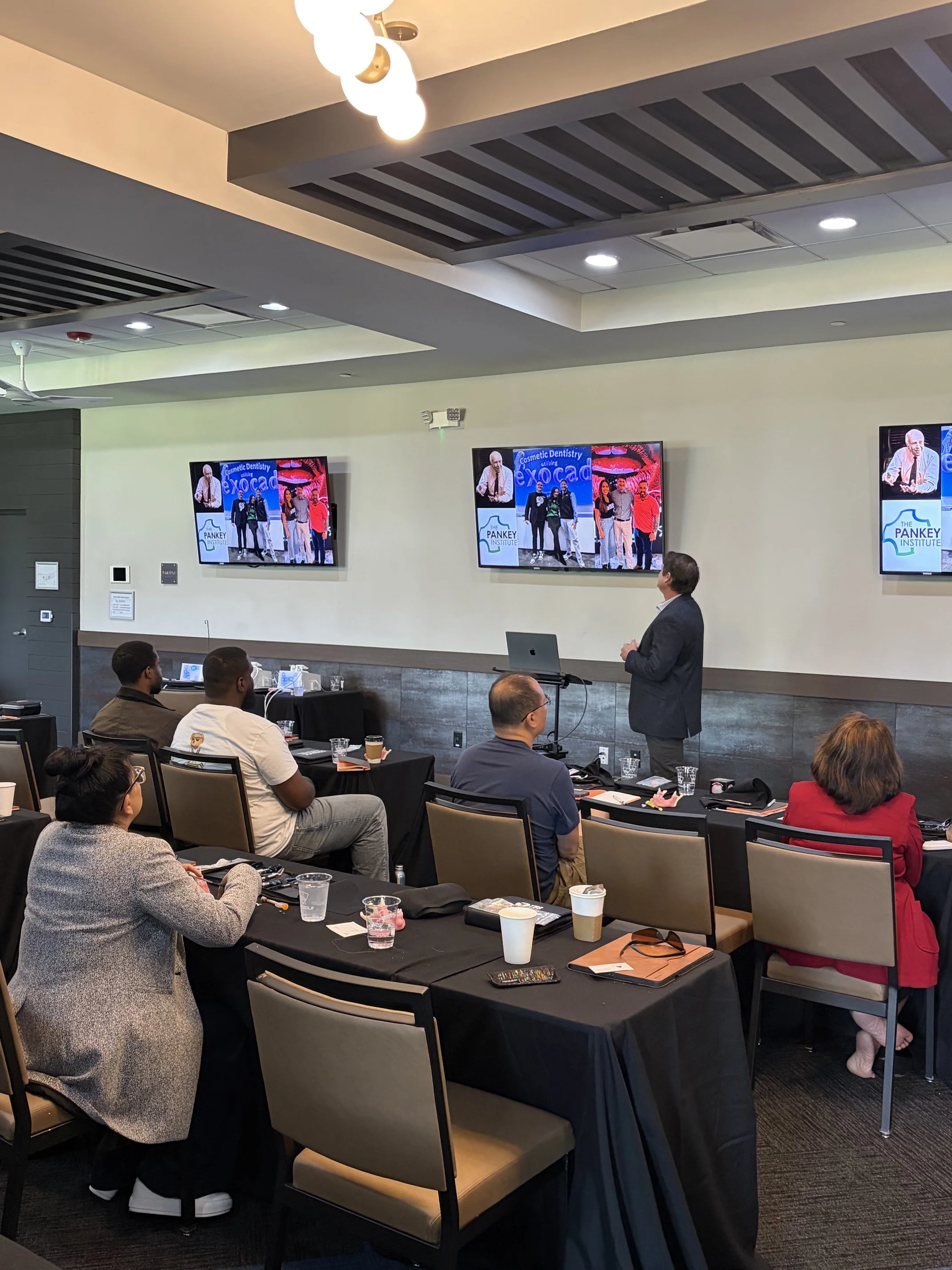 A group of people seated at tables in a conference room listening to a speaker standing near a presentation screen. The screen displays a dental themed graphic and the text 'Cosmetic Dentistry Village Exocad' and 'The Pankey Institute.' The room has a modern ceiling design with lights and fans.