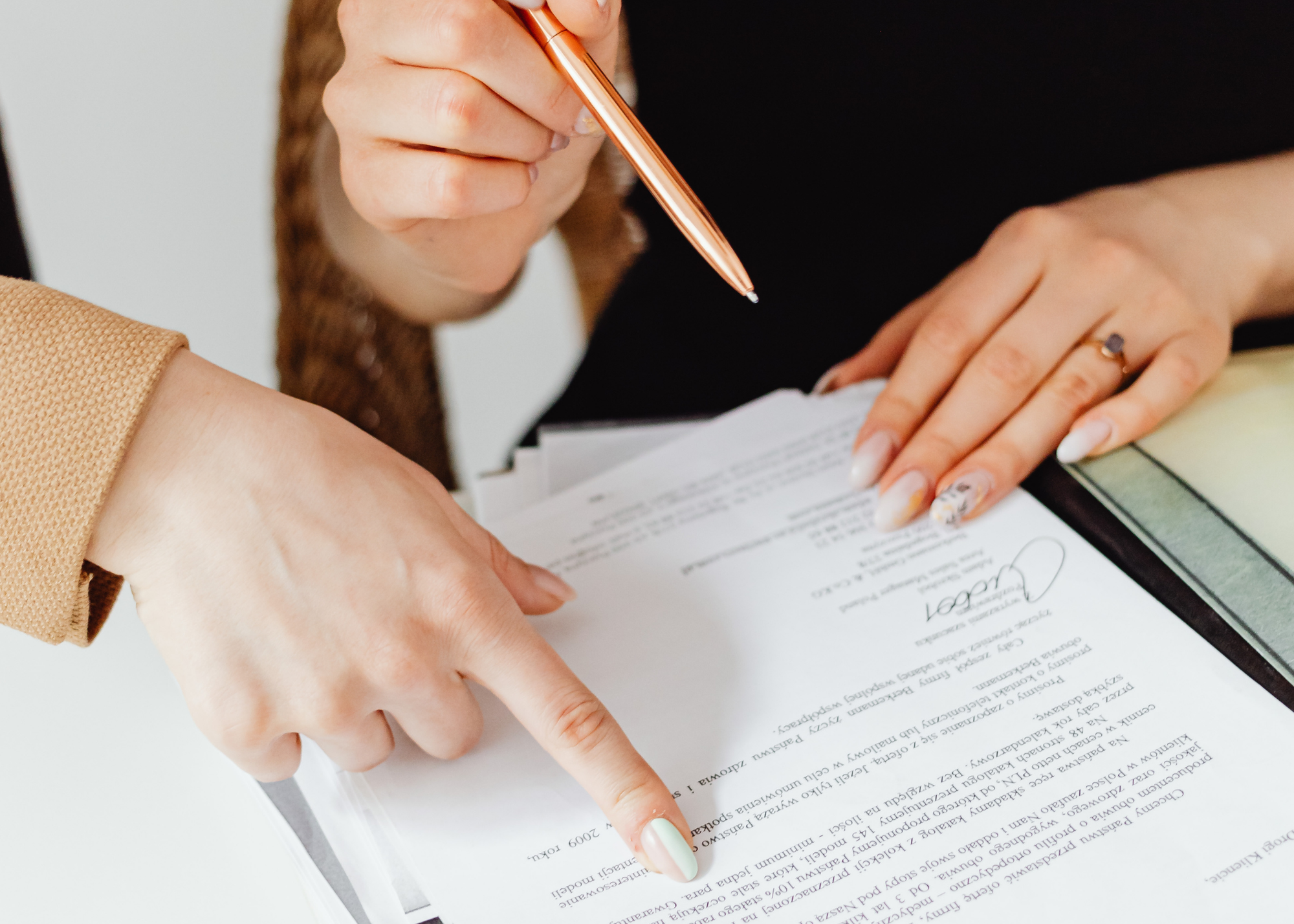 Person wearing black shirt holding gold pen pointed at paper with text and signature and another with orange blazer sleeve showing pointing at document