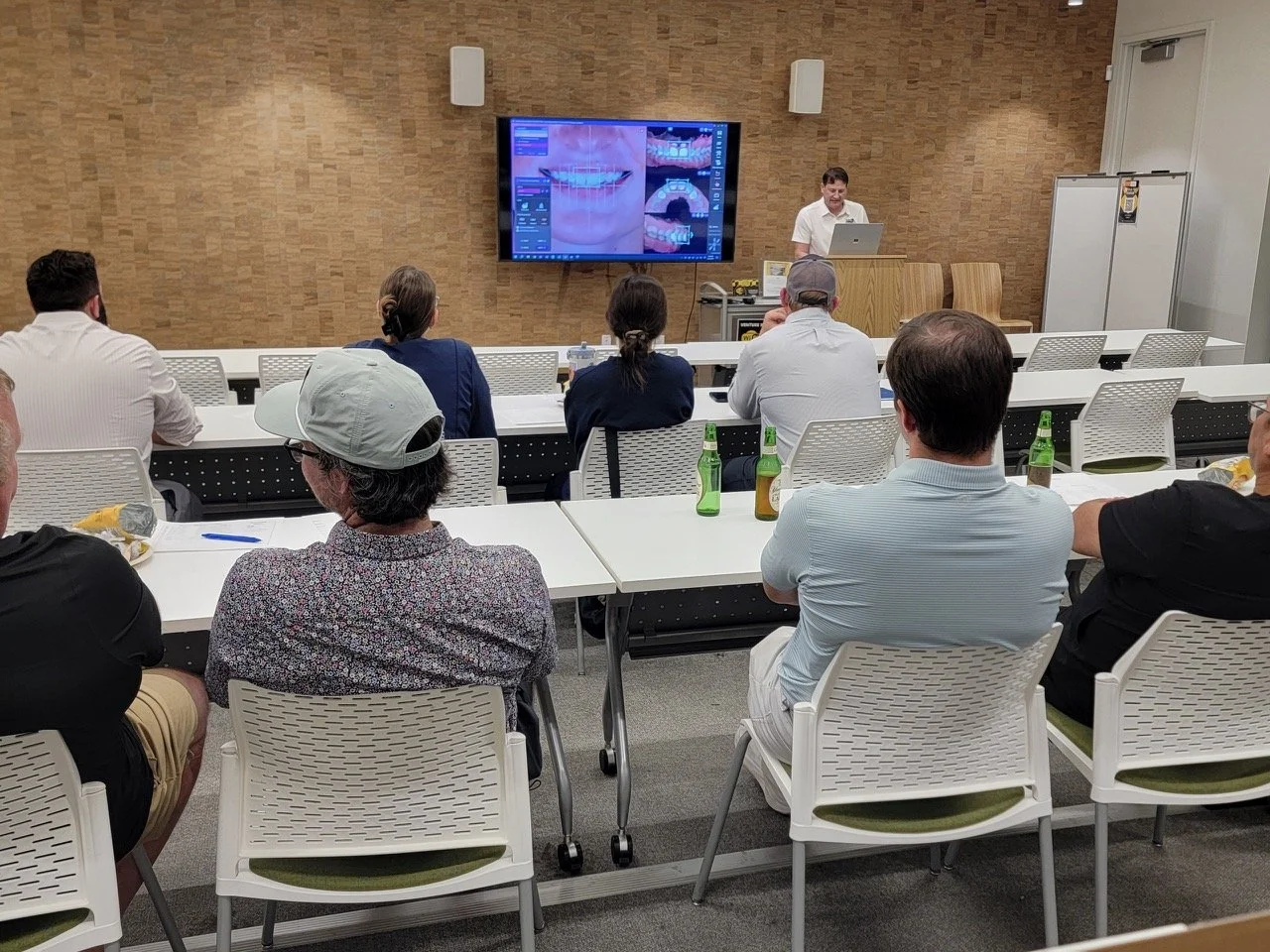 A man is giving a presentation in a conference room with an audience watching a slide showing dental images on a large screen.