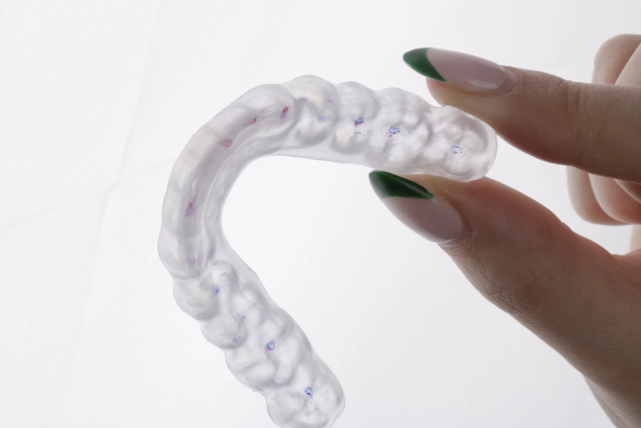 Close-up of a hand holding a clear, flexible dental aligner with small colored specks, showing green and beige French manicured nails.
