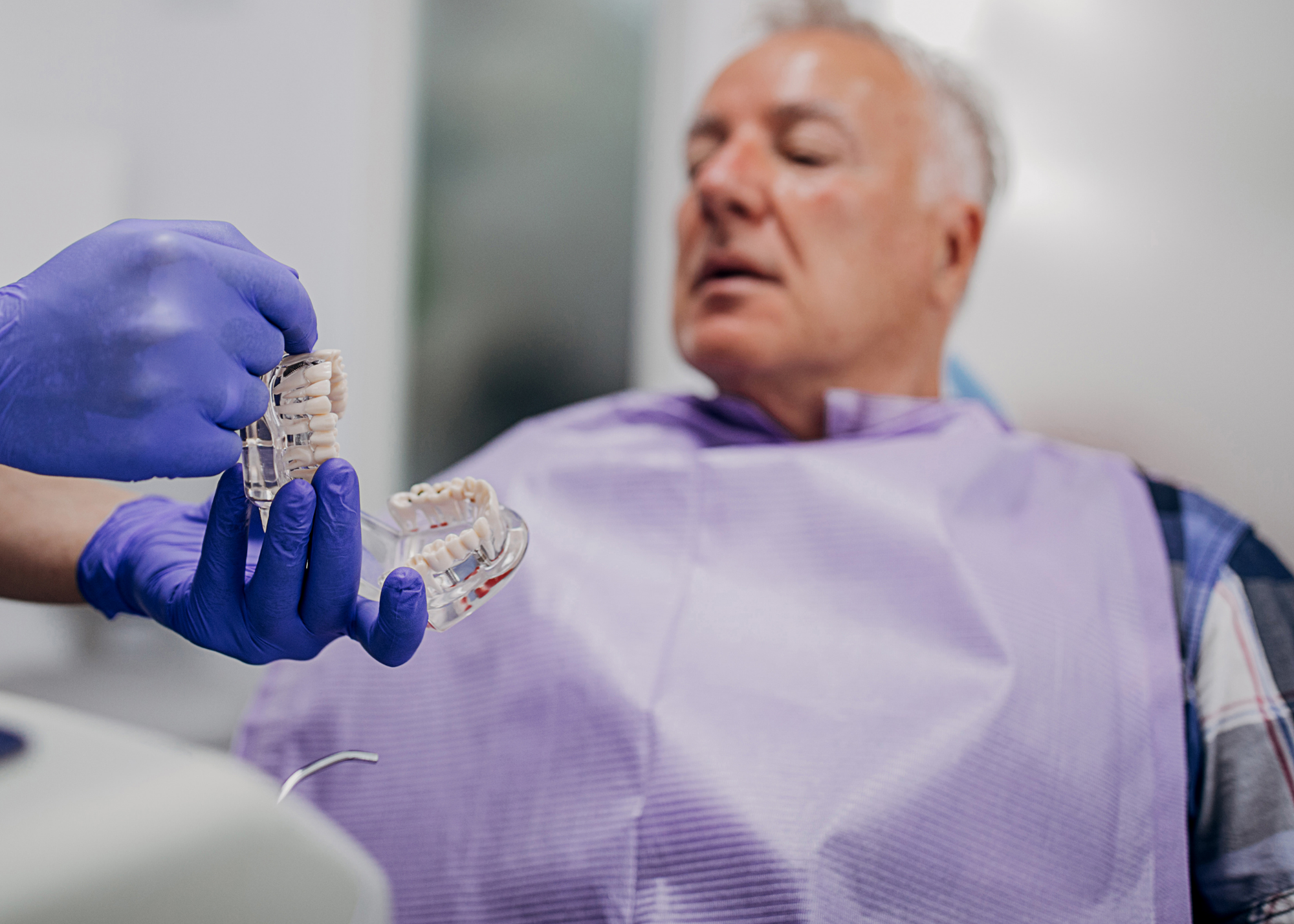 man sitting in dental chair with purple dental bib on being shown dental model held by purple gloves in a dental office.