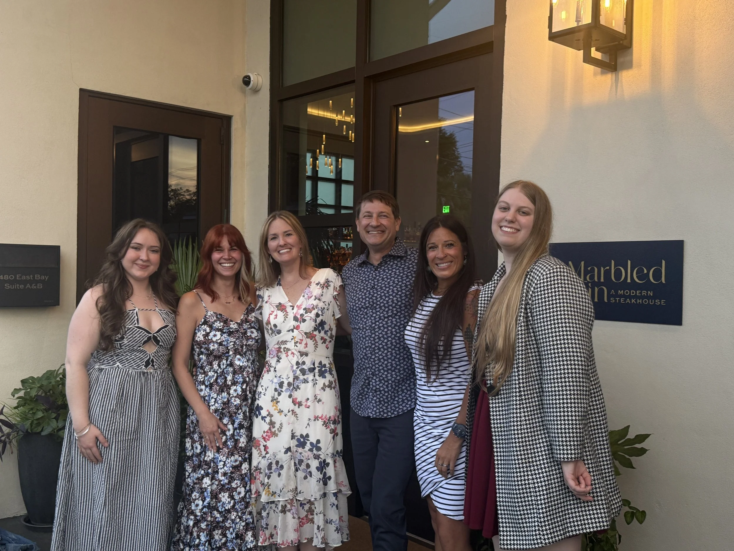 Group of six smiling people standing outside a modern restaurant with large windows and a sign to the right that reads "Marbled..."