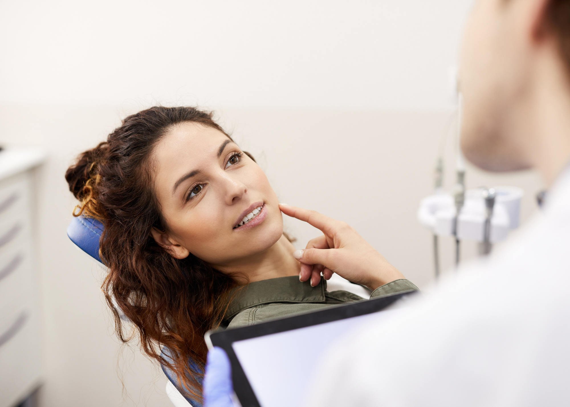 Woman with long dark hair and brown eyes reclined in dental chair pointing at the side of her jaw