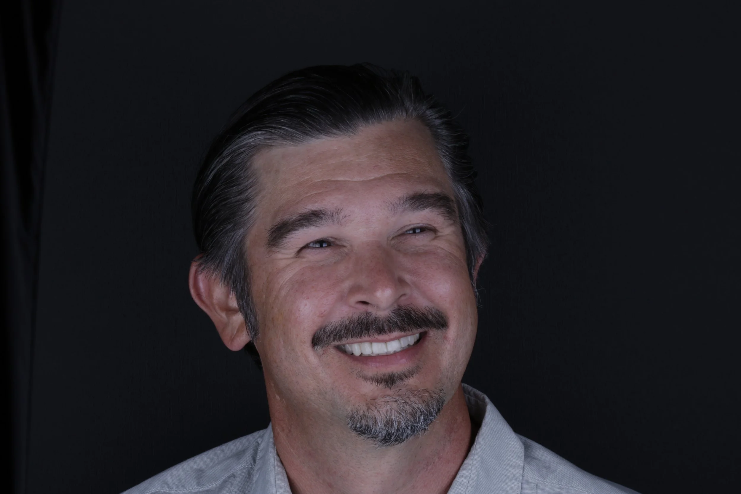 Close-up photo of a middle-aged man with dark hair graying at the temples, smiling, wearing a light-colored shirt against a black background.