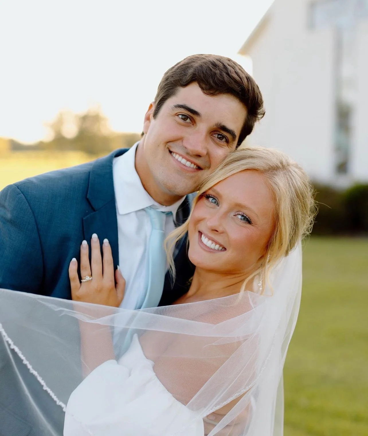A smiling bride and groom embrace outdoors during daytime, with the groom in a blue suit and the bride in a white wedding dress and veil.