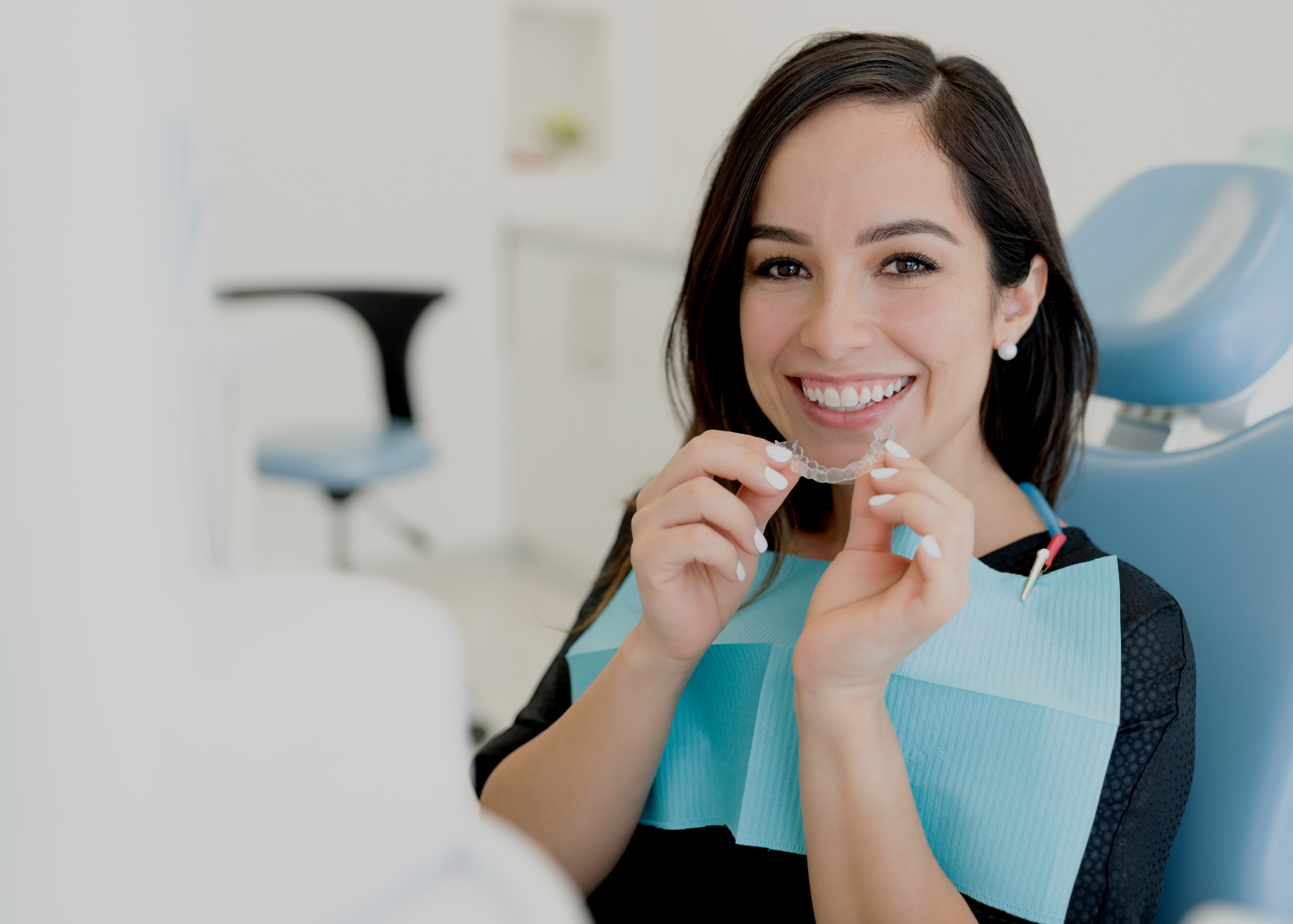 A woman smiling in dental office with a blue dental bib on and holding a clear dental aligner in her hand up to her mouth.