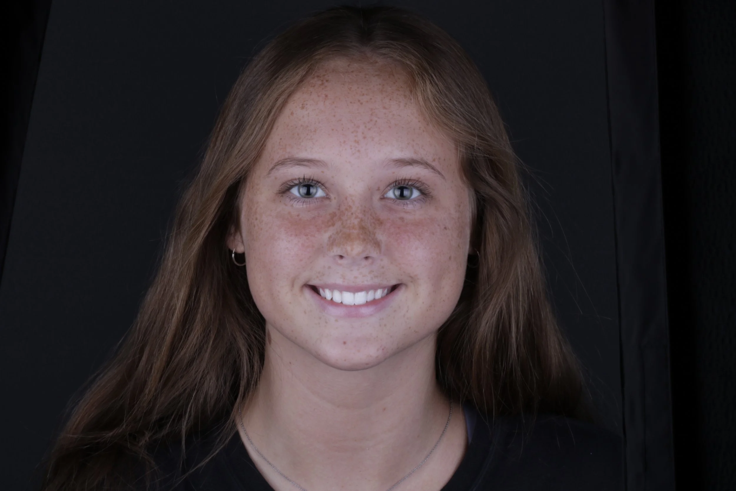 Close-up of a young woman with red hair, freckles, and blue eyes smiling, against a black background.
