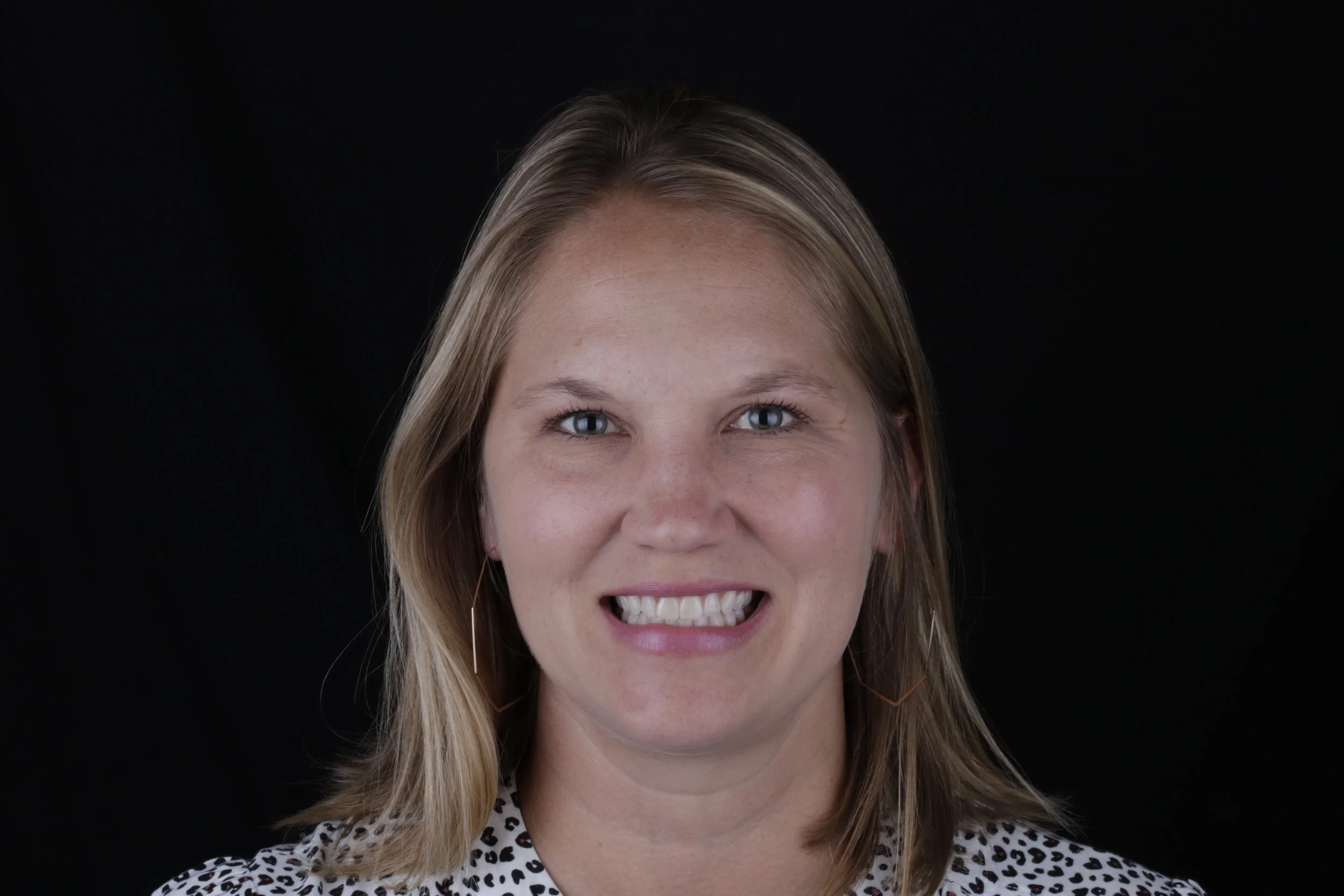 A woman with blonde hair smiling, wearing a leopard print top, against a black background.