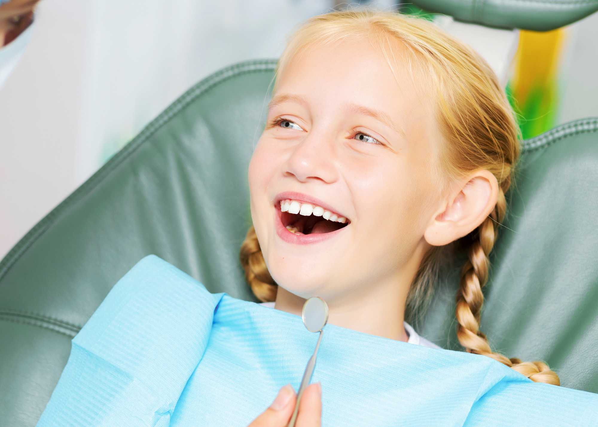 A young girl with blonde hair in braids lying on a dental chair, smiling happily, with a dental mirror in her hand, in a dentist's office.