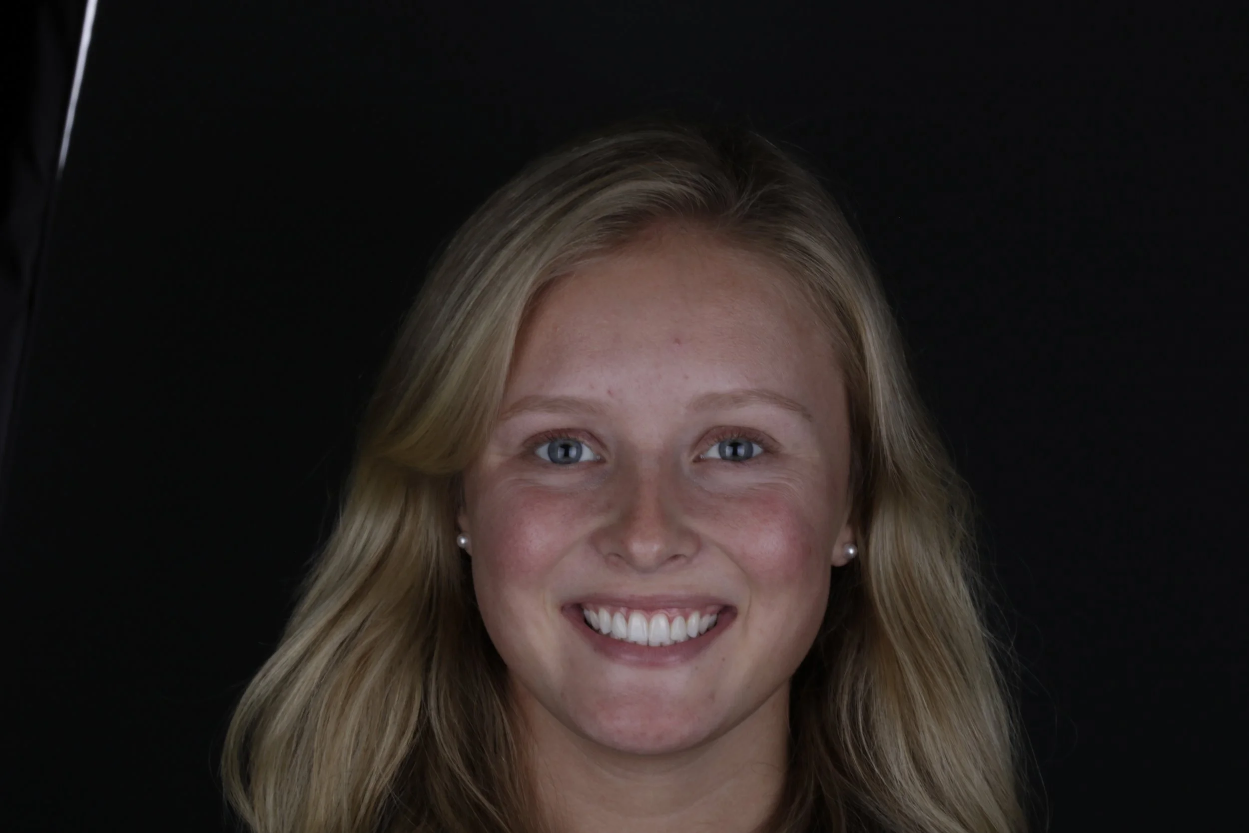 Close-up of a smiling woman with blonde hair and blue eyes, against a black background.
