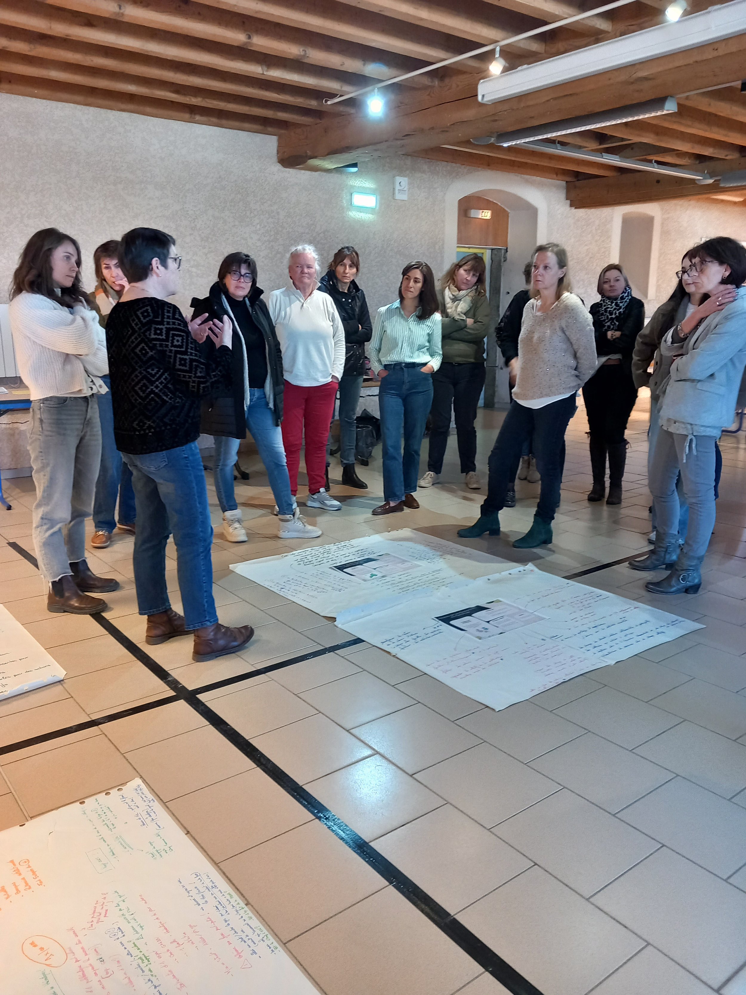 Groupe de personnes discutant autour de grandes feuilles de papier posées sur le sol dans une pièce avec plafond en bois.