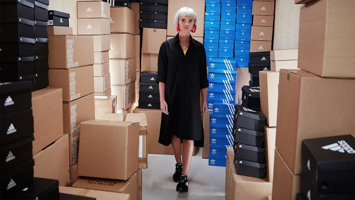 Person walking through a room filled with stacked cardboard boxes and shoe boxes, including Adidas branded ones; wearing black outfit and red earrings.