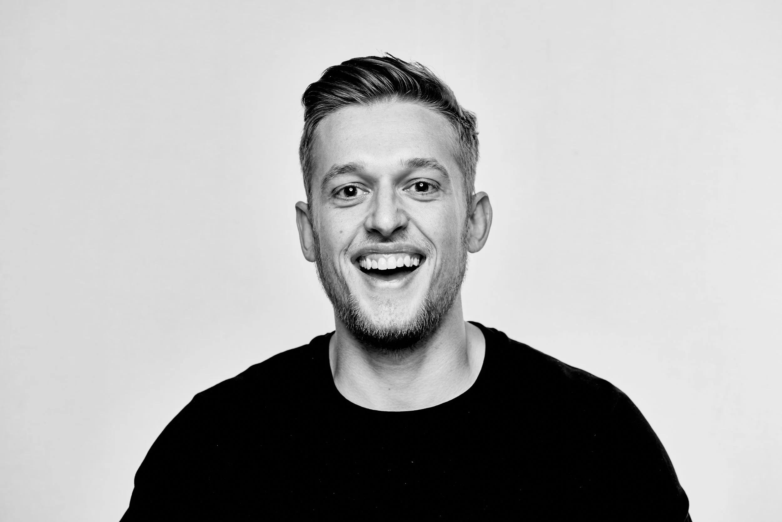 Black and white portrait of a smiling man in a black shirt against a plain background.