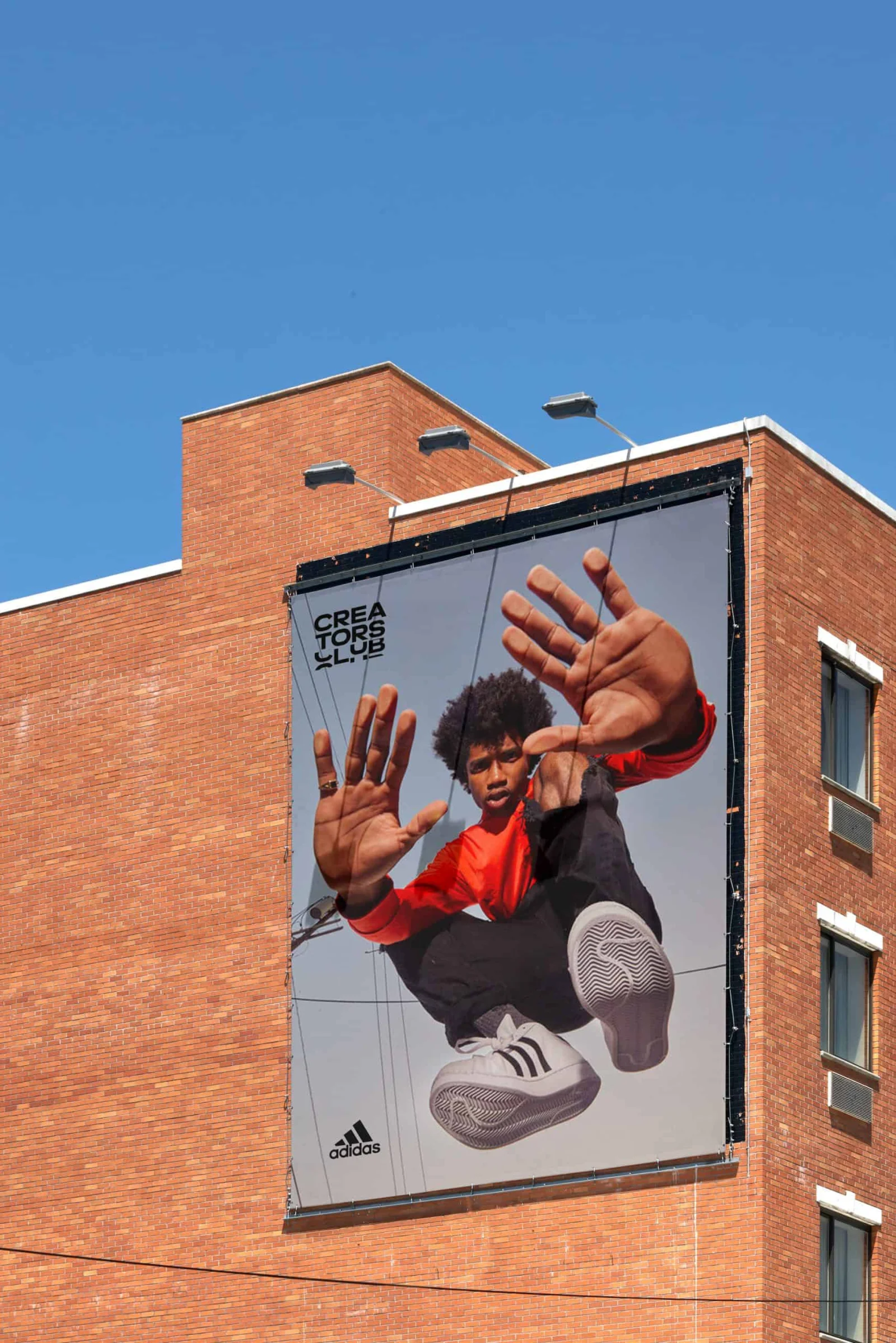 Large Adidas billboard on a brick building featuring a person in a dynamic pose, wearing a red shirt and Adidas shoes, with "Creators Club" text.