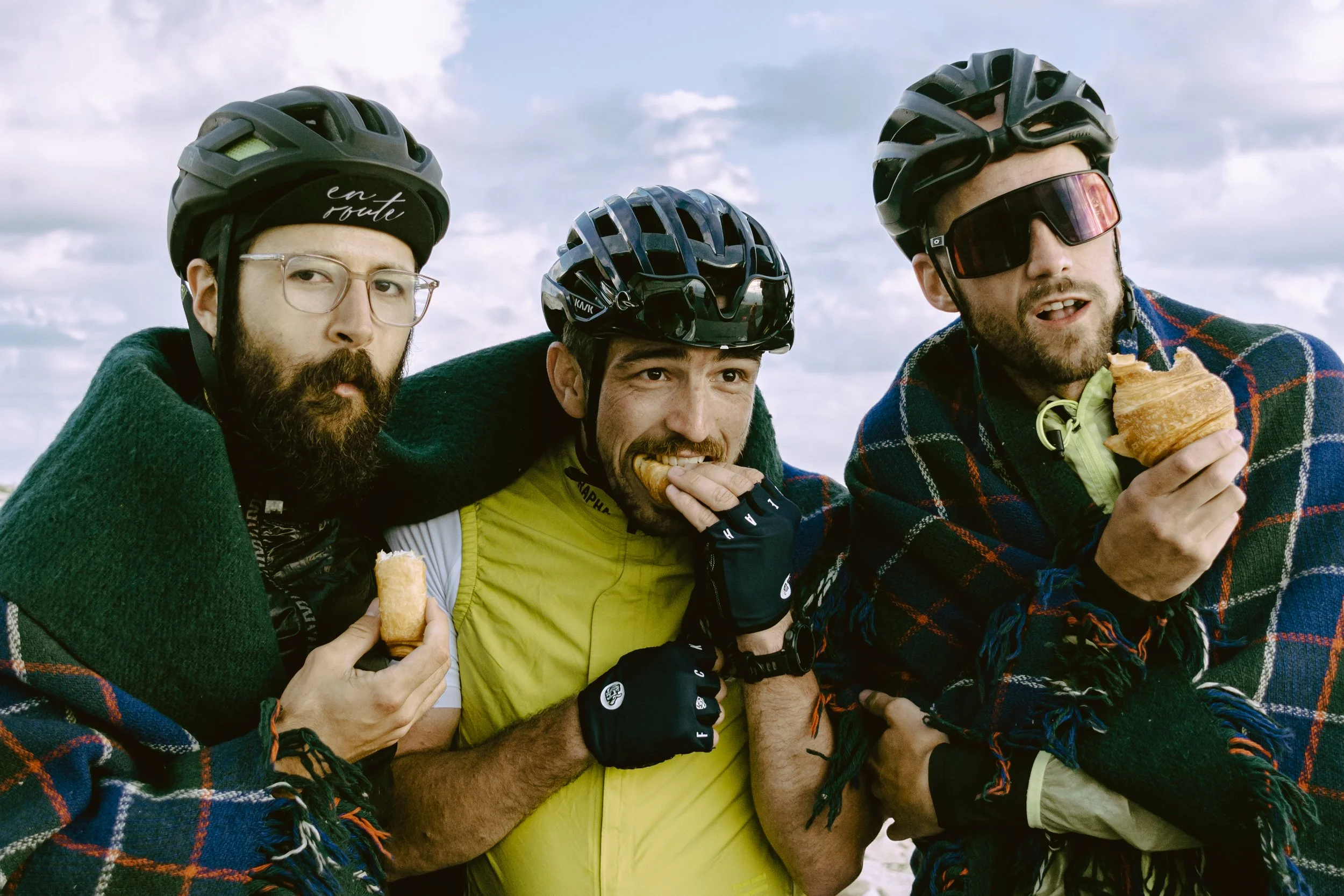 Three cyclists wearing helmets and glasses, wrapped in blankets, eating pastries outdoors under a cloudy sky.