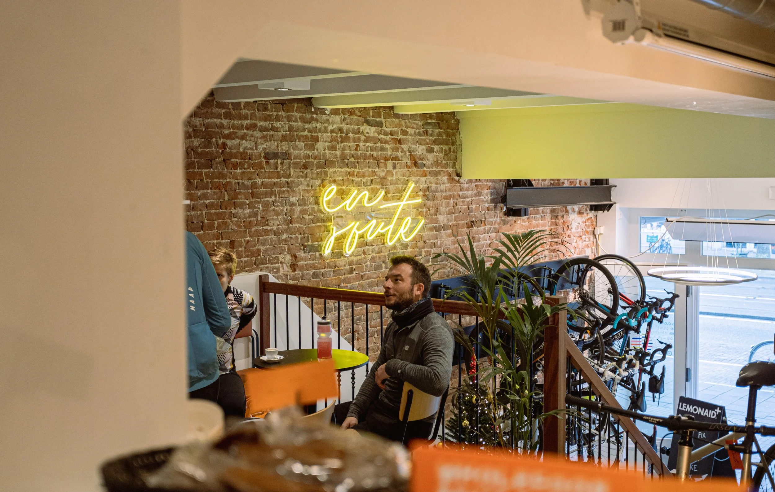 Interior of a cafe with a brick wall, neon sign 'en route', and hanging bicycles. People are seated, conversing at small tables. Plants decorate the space, and there's a large window visible. A LemonAid sign is seen near bicycles.