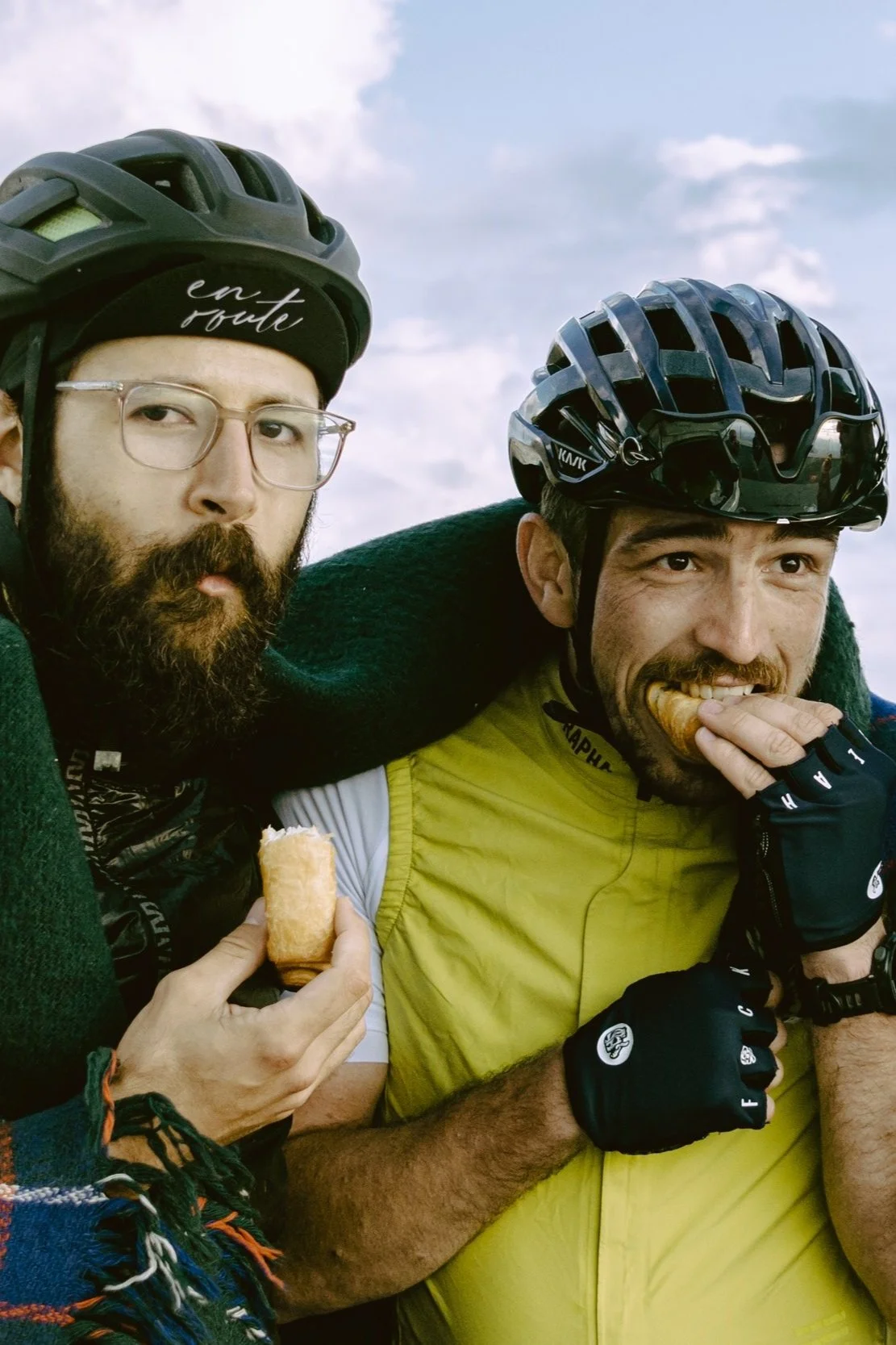 Two cyclists with helmets, eating snacks and wearing outdoor gear, one in a green scarf and the other in a yellow vest.