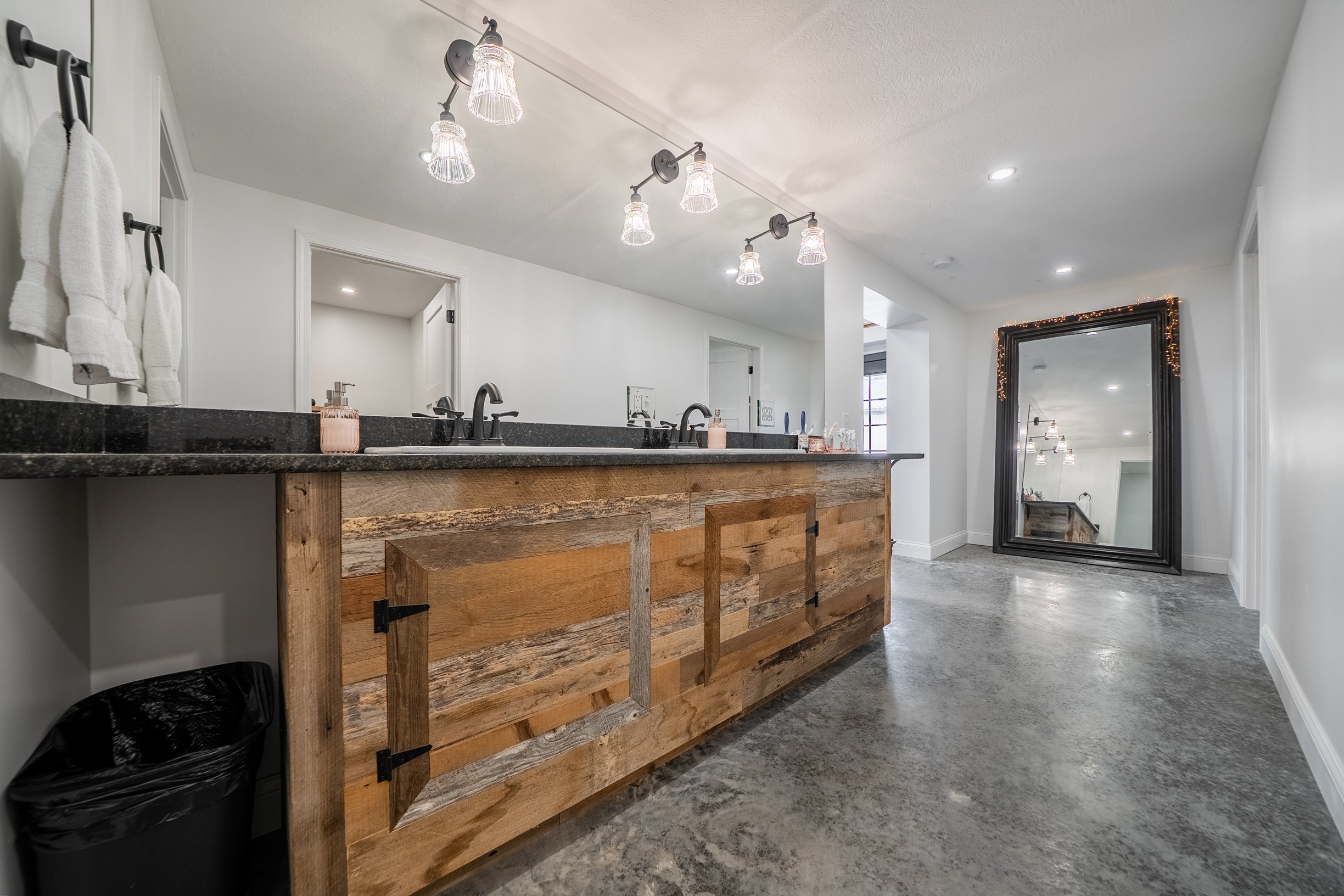 Bathroom with white walls, a large mirror decorated with string lights, a black granite countertop with two sinks, and a rustic wooden vanity. There are white towels hanging on the wall, a soap dispenser, and a trash bin. The ceiling has exposed ligh