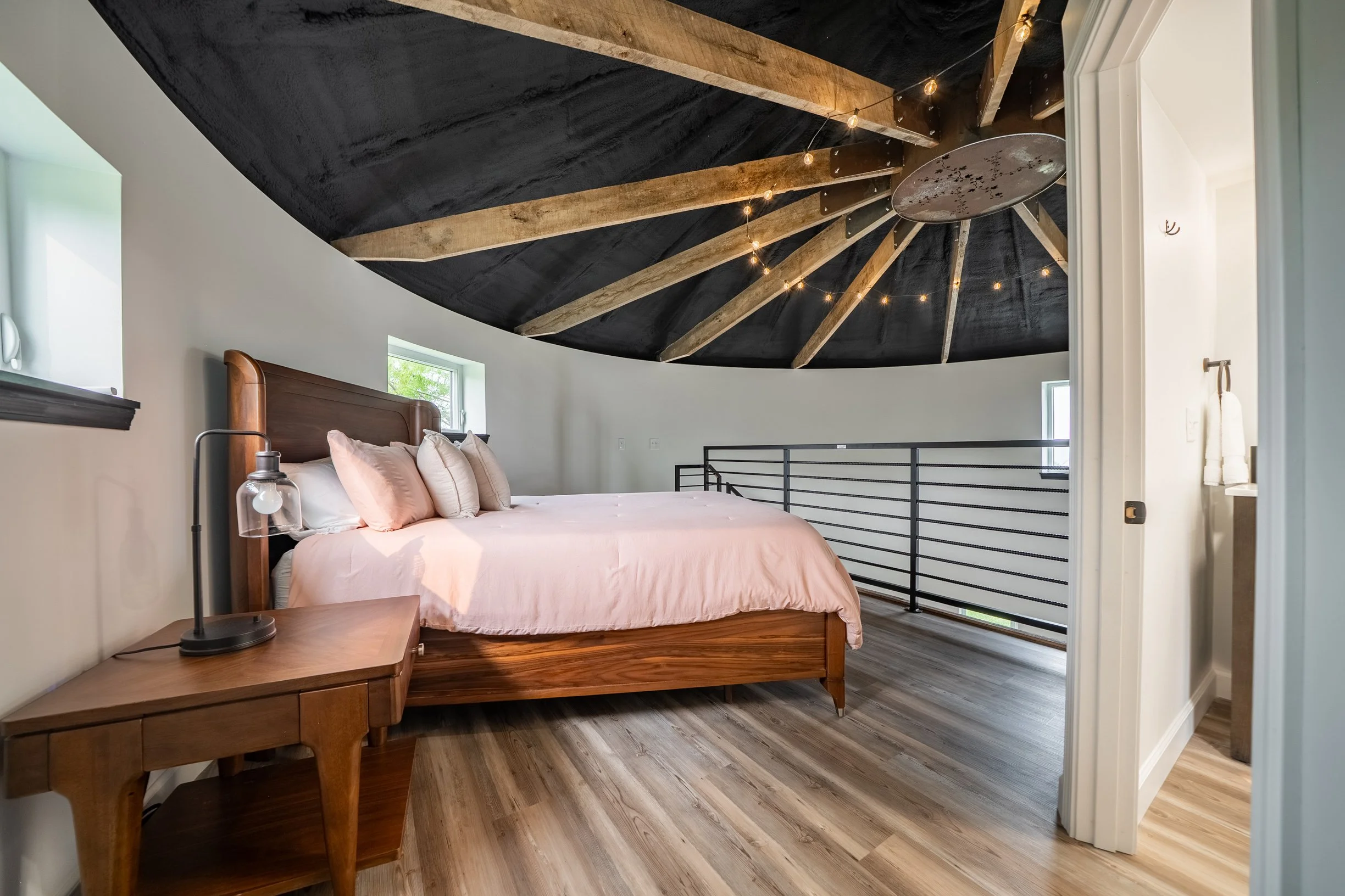 A bedroom with a wooden bed frame, pink bedding, multiple pillows, a nightstand with a modern lamp, small windows with green trees outside, dark wood flooring, and a vaulted ceiling with black paint and wooden beams with string lights.
