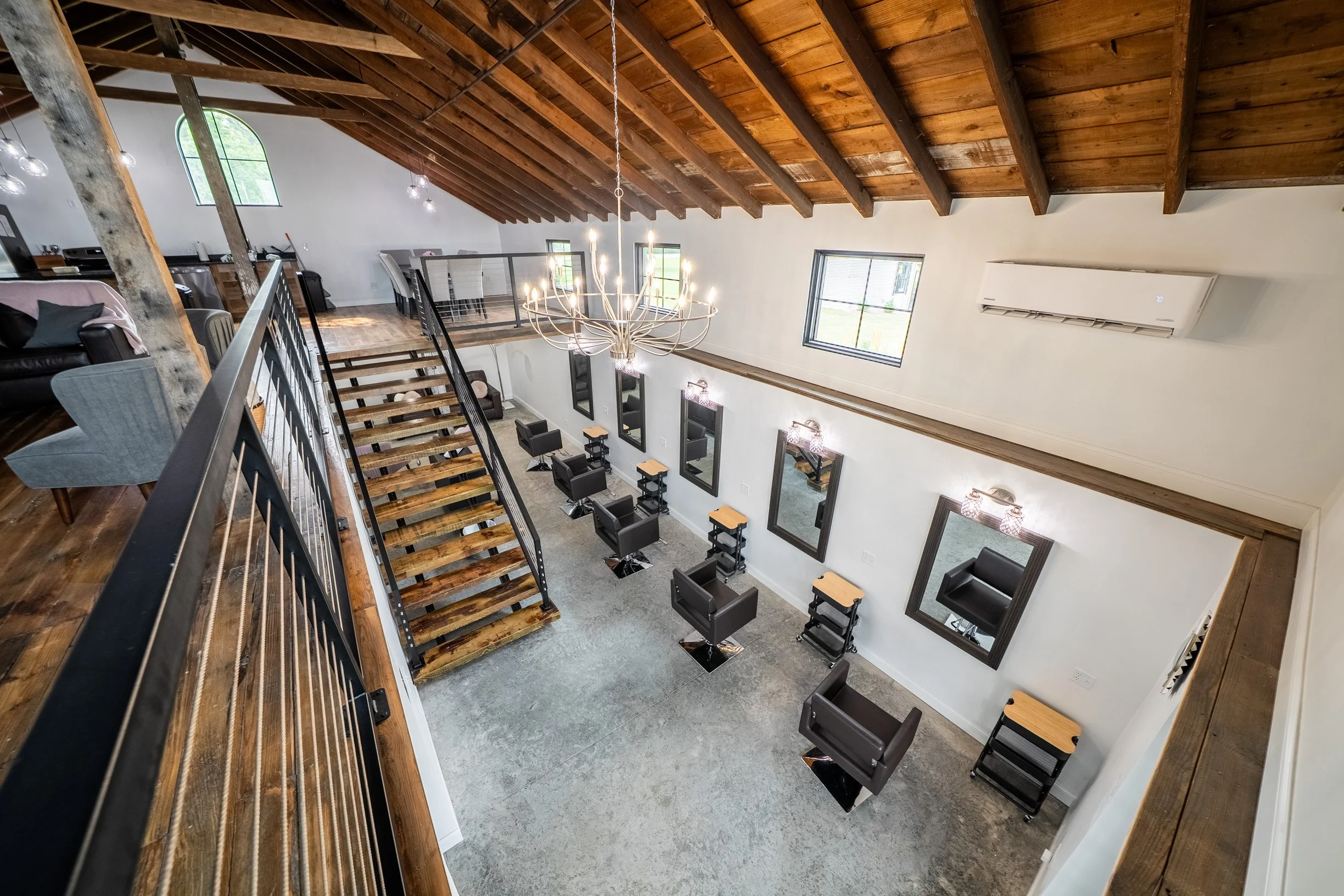 A spacious indoor area with a high wooden, sloped ceiling, large windows, and modern black salon chairs aligned against the white walls. There is a staircase with wooden steps and black railings leading to a mezzanine level, where a dining area with a table and chairs is visible. Elegant light fixtures hang from the ceiling, including a large chandelier. An air conditioning unit is mounted on the wall.