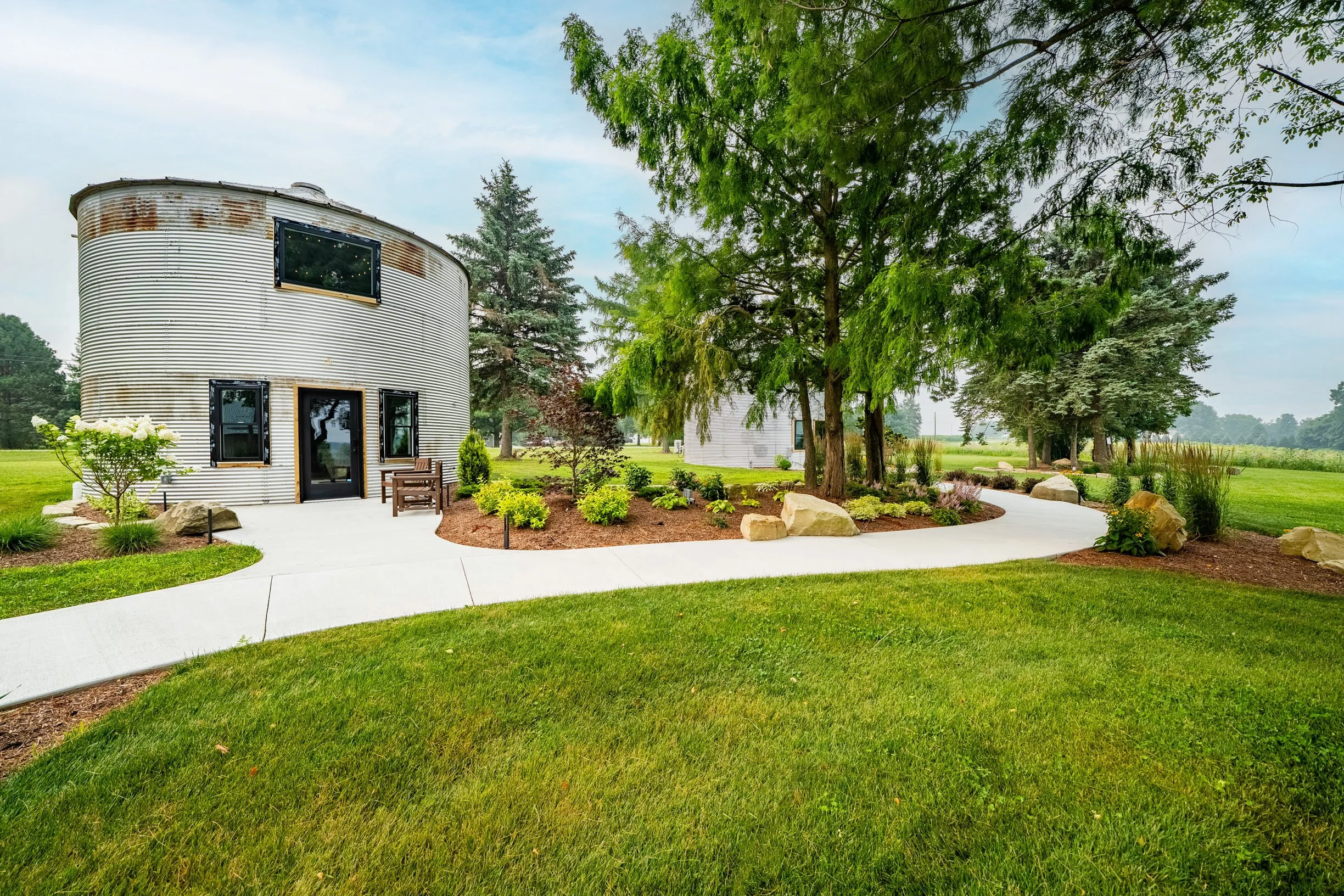 A modern, cylindrical silver building with black-framed windows and a black door, surrounded by a landscaped garden with green grass, bushes, trees, and large rocks. A concrete pathway runs through the garden under a blue sky with some clouds.