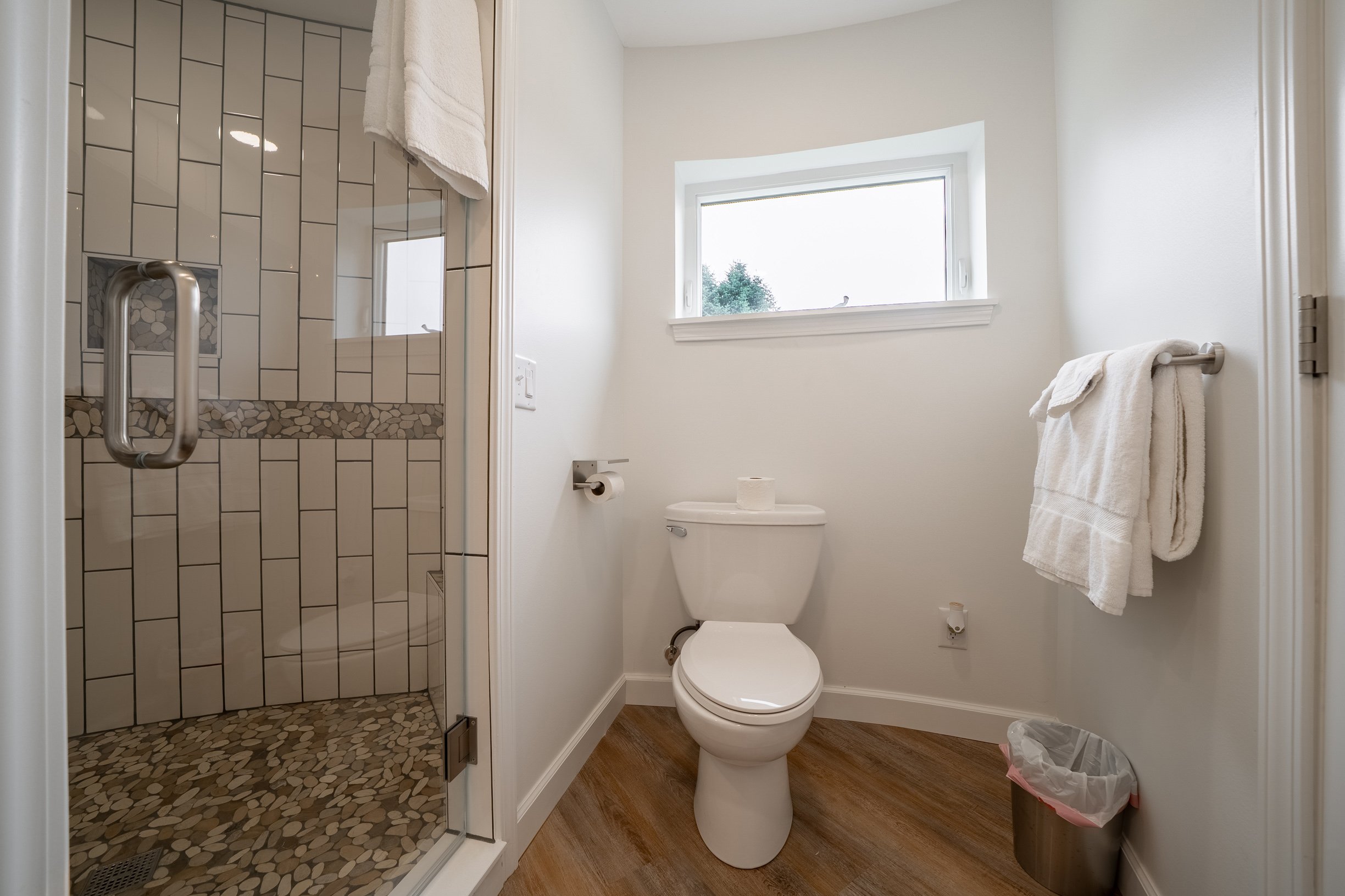 A small bathroom with a shower on the left with glass door, white tile walls, and pebble accent tiles; a white toilet and a window above it; white walls, a towel rack with towels, and a trash can.