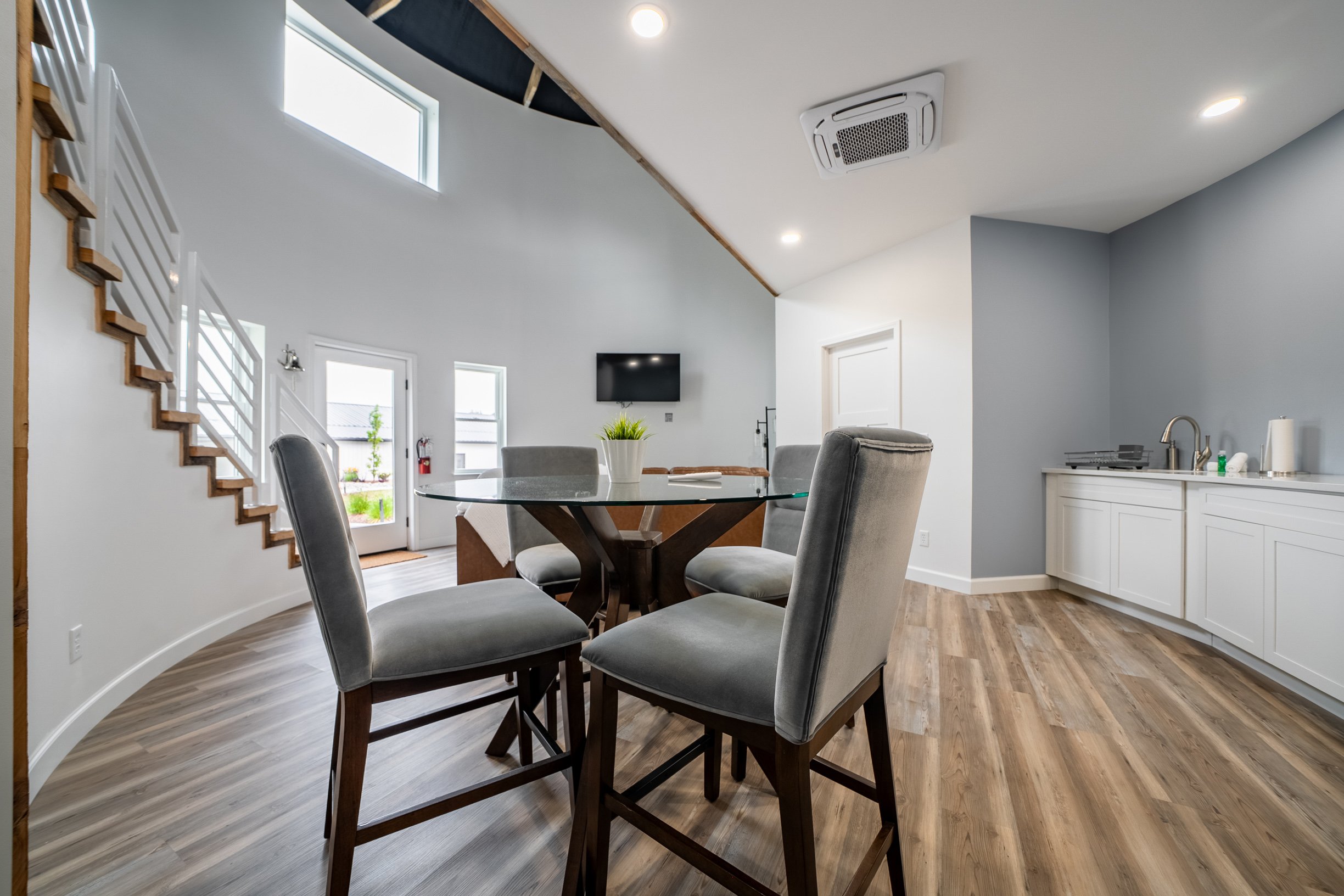 Modern dining area with a round glass table surrounded by six gray upholstered chairs in a bright room with wood flooring, a small kitchen area, a wall-mounted TV, and large windows letting in natural light.