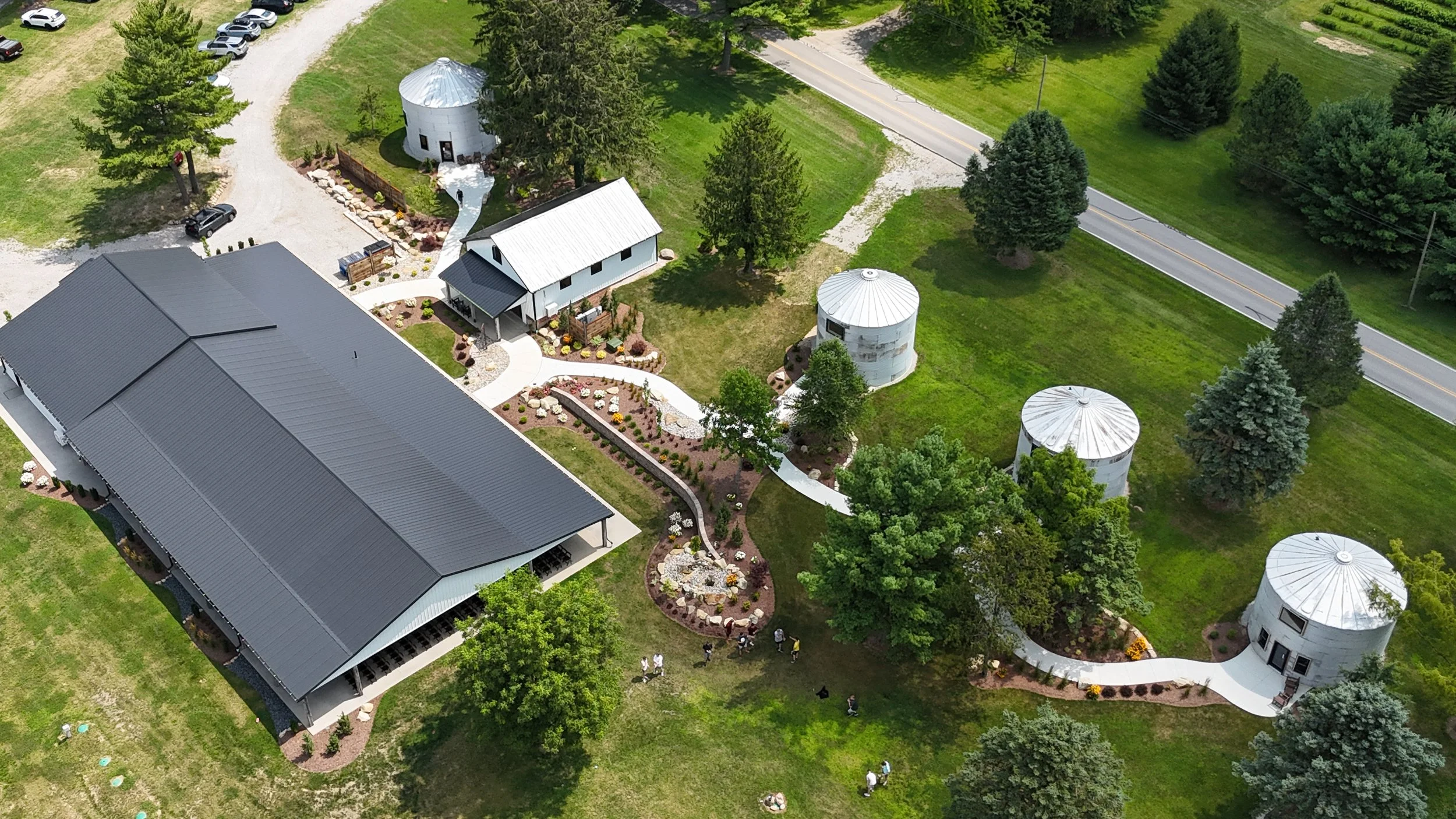 An aerial view of a rural property with a large building with a metal roof, a smaller white barn-style building, and several silos. The property features landscaped gardens, curved pathways, and numerous trees, with a nearby road and green fields.