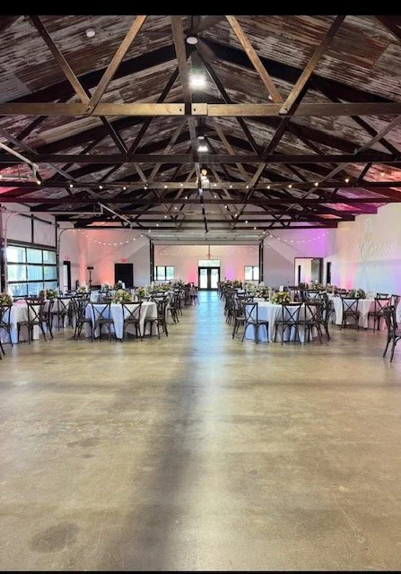 Event space with round tables covered in white tablecloths, decorated with floral centerpieces, in a large industrial-style hall with a high wooden ceiling, string lights, and a hardwood floor.