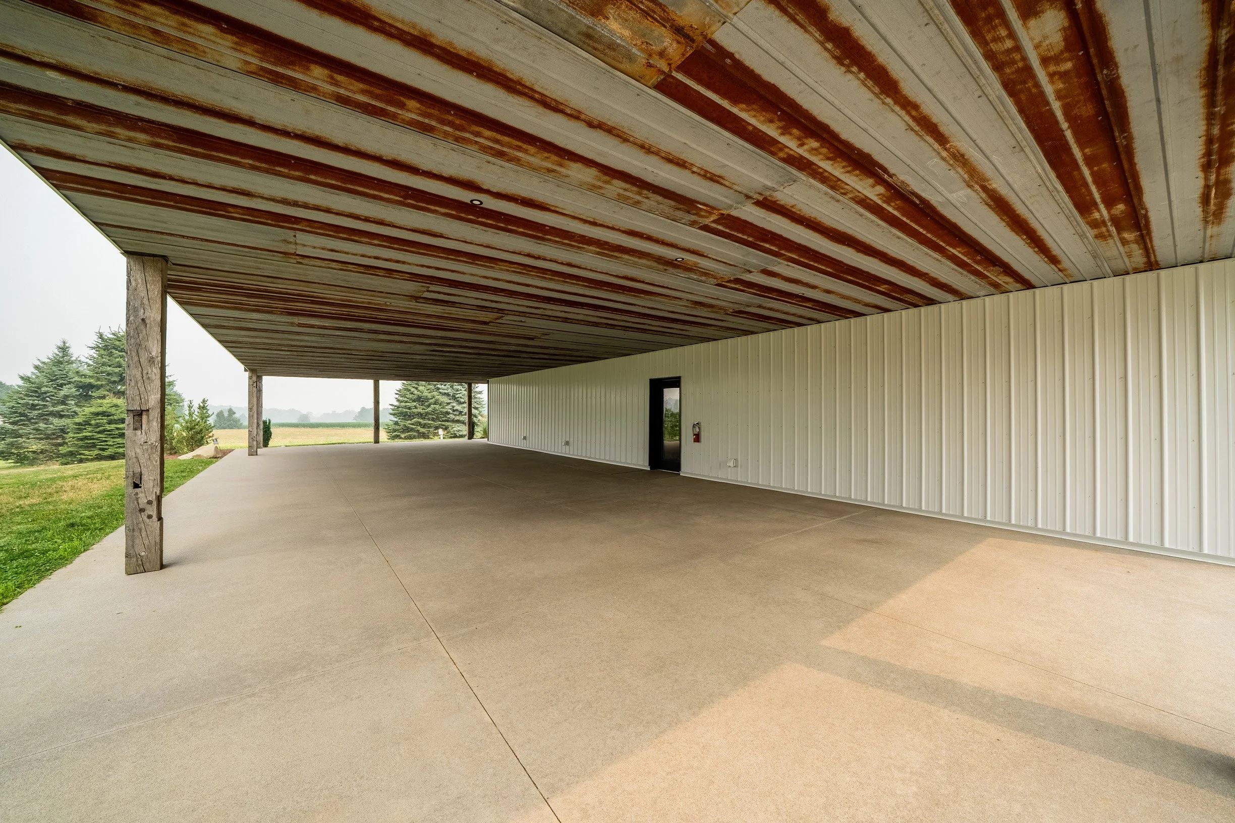A covered outdoor patio area with a concrete floor, supported by wooden posts, with a rusted corrugated metal roof and beige metal siding on the building wall. In the background, there are green trees and a cloudy sky.
