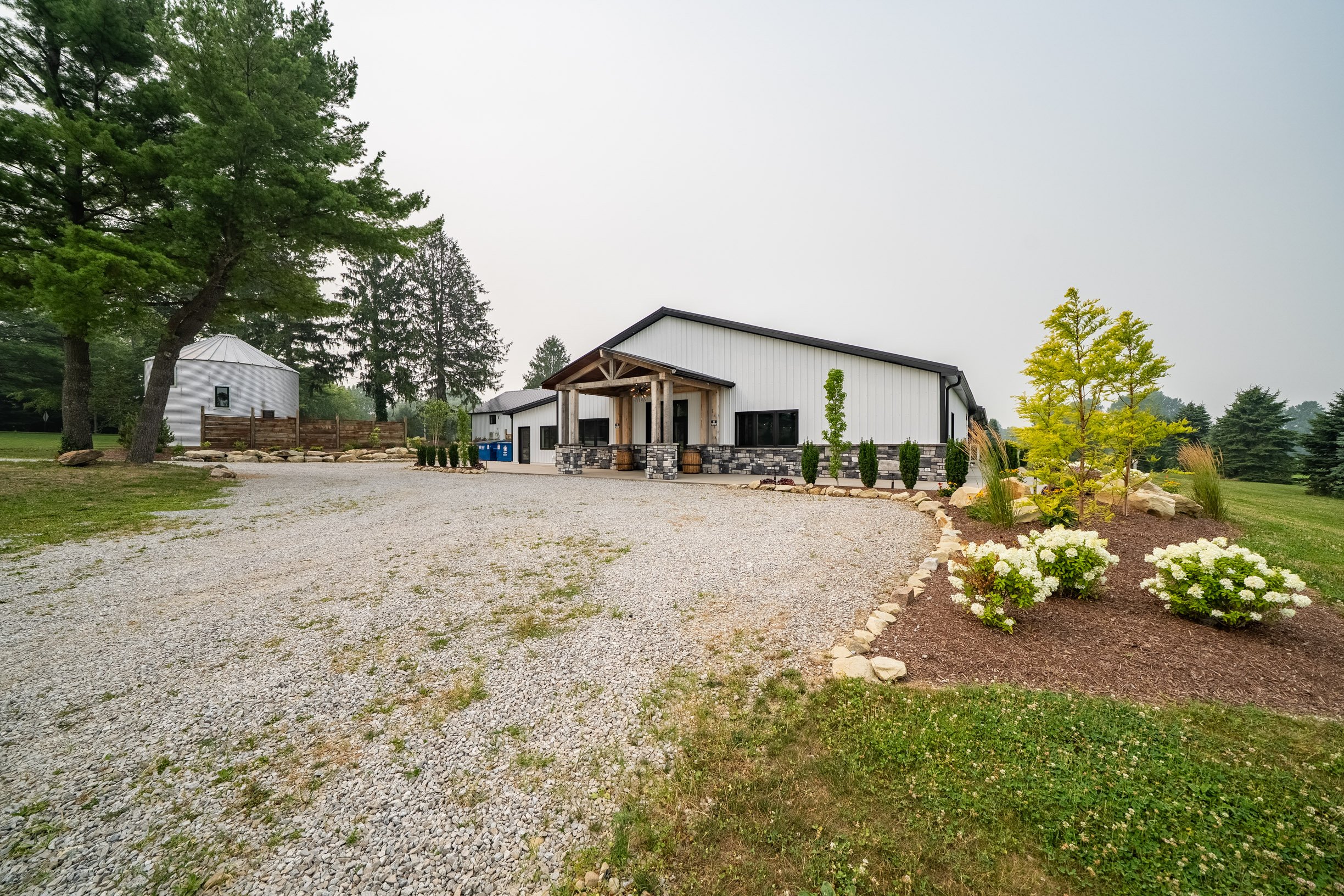 A white modern barn-style building with black roofline and stone foundation, surrounded by landscaped plants and trees, with a gravel driveway and a green lawn in the background.