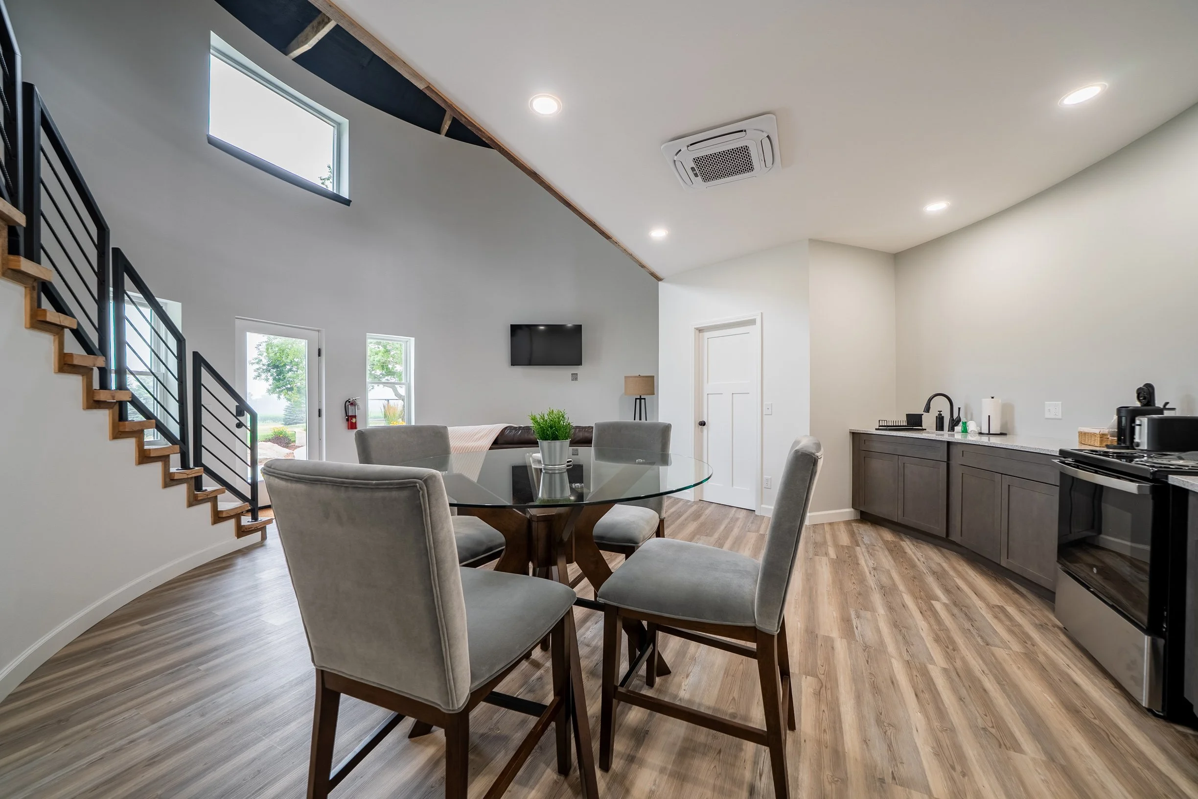 Interior of a modern home with a dining area and kitchen. Features include a round glass dining table with six beige upholstered chairs, a kitchen with dark cabinets, stainless steel appliances, a sink, and countertop items. Natural light enters through large windows, and there is a staircase with wooden steps and black railings.
