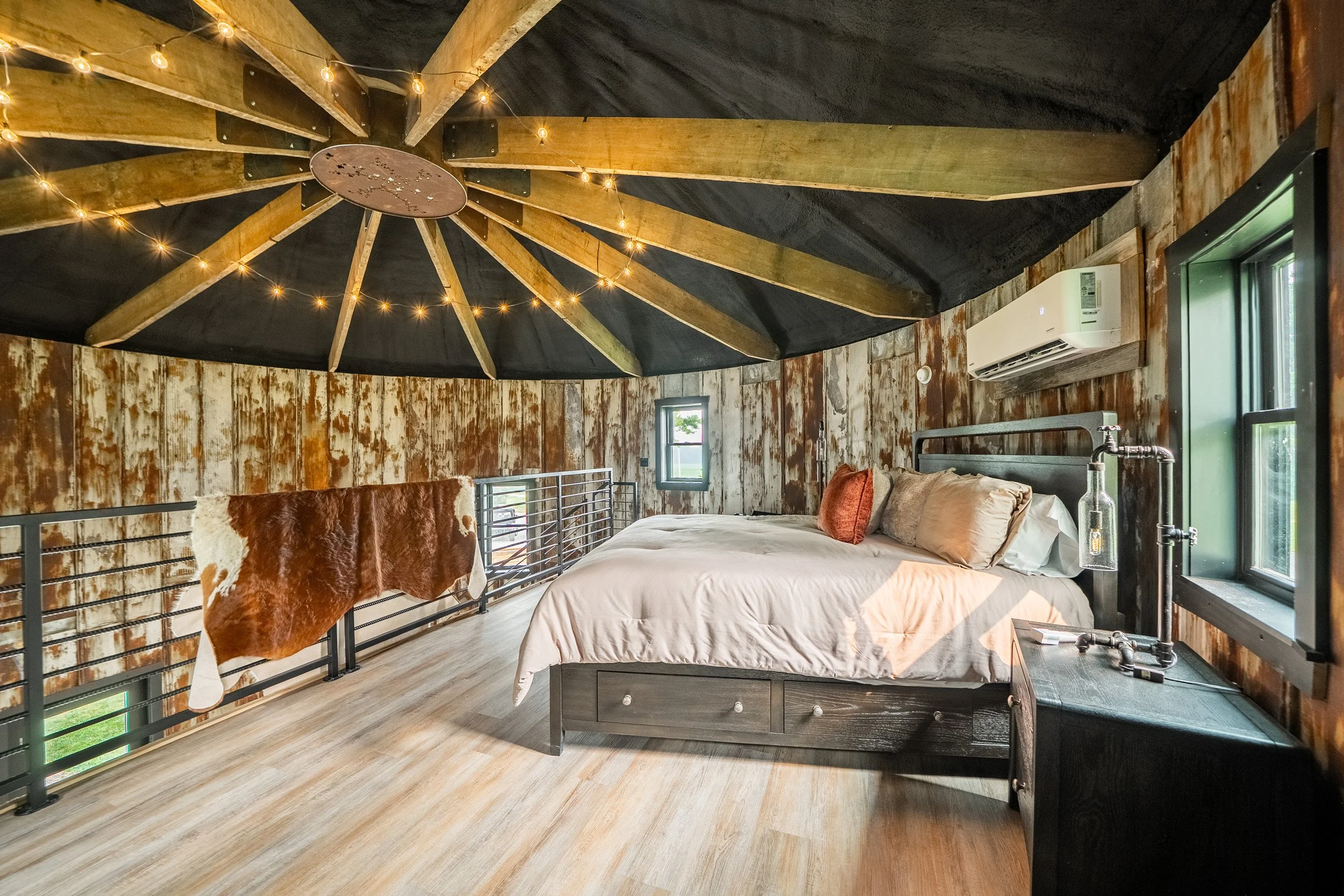 Bedroom with rustic wood walls, a black bed frame, and a large bed with beige bedding and pillows. A cowhide blanket is draped over a black metal railing, and string lights hang from the ceiling. There are two small windows and a modern air conditioner on the wall.