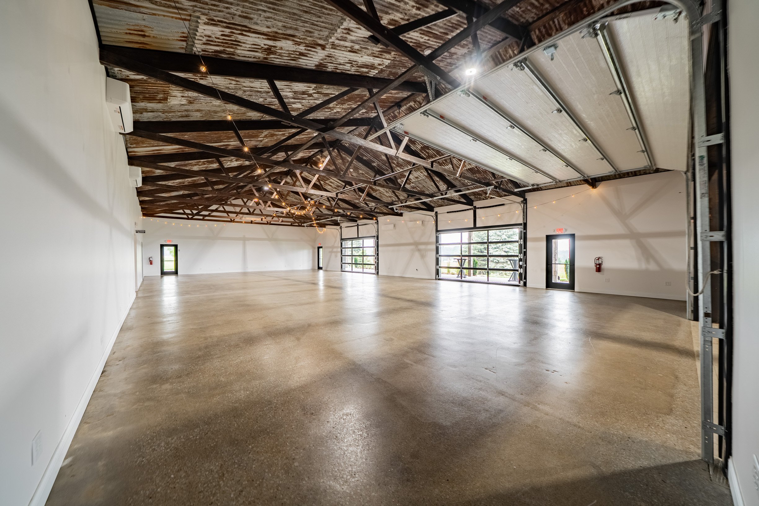 Interior view of an empty modern industrial-style event or meeting space with high ceilings, exposed wooden beams, string lights, large glass garage doors, and polished concrete floors.