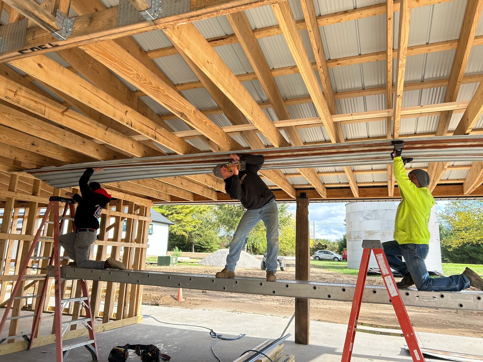 Three construction workers installing metal sheets on a wooden structure roof, using ladders and scaffolding, with outdoor scenery in the background.