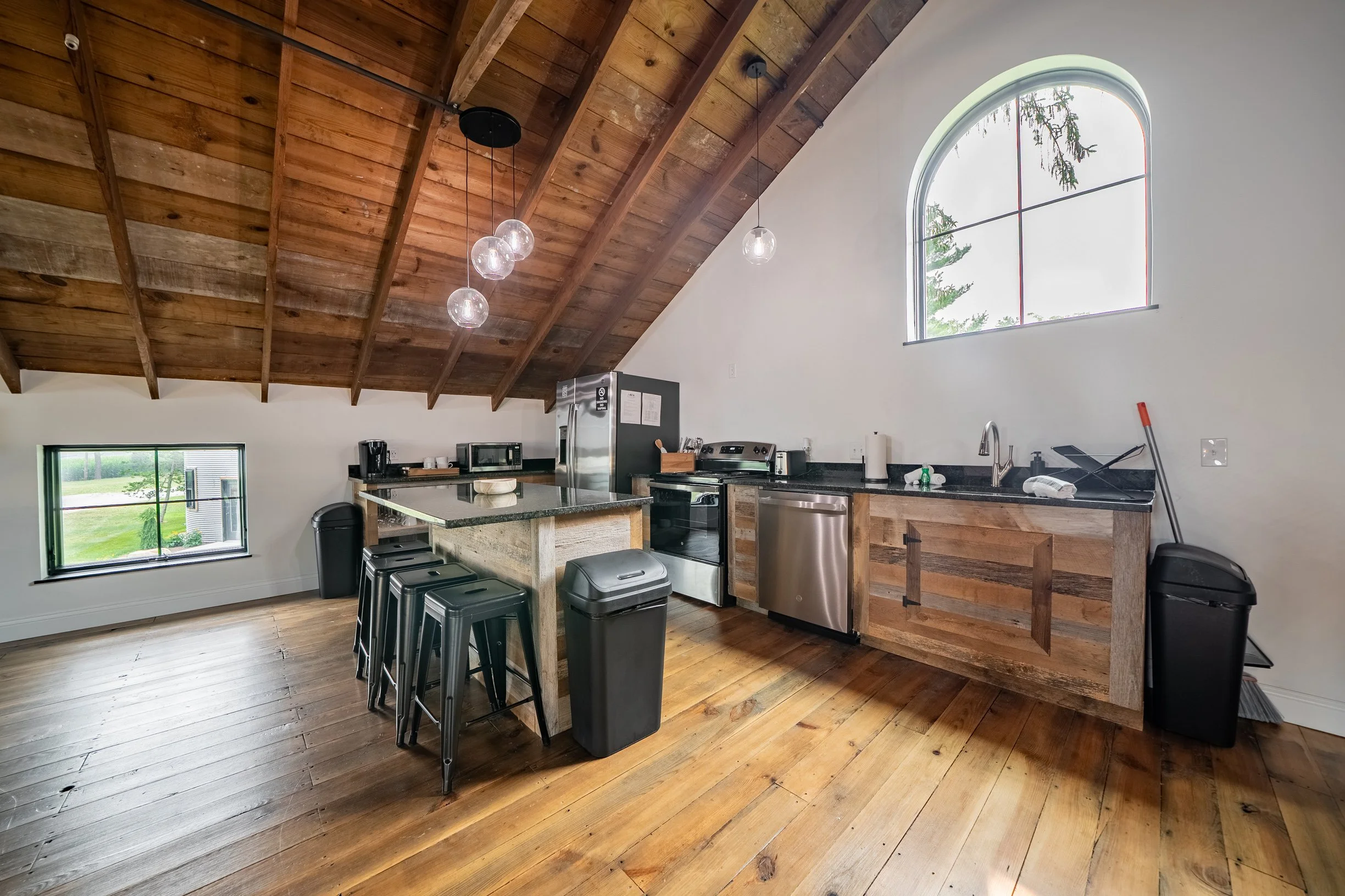 Kitchen with wooden vaulted ceiling, hanging pendant lights, granite countertops, stainless steel appliances, and a large arched window with a view of trees outside.