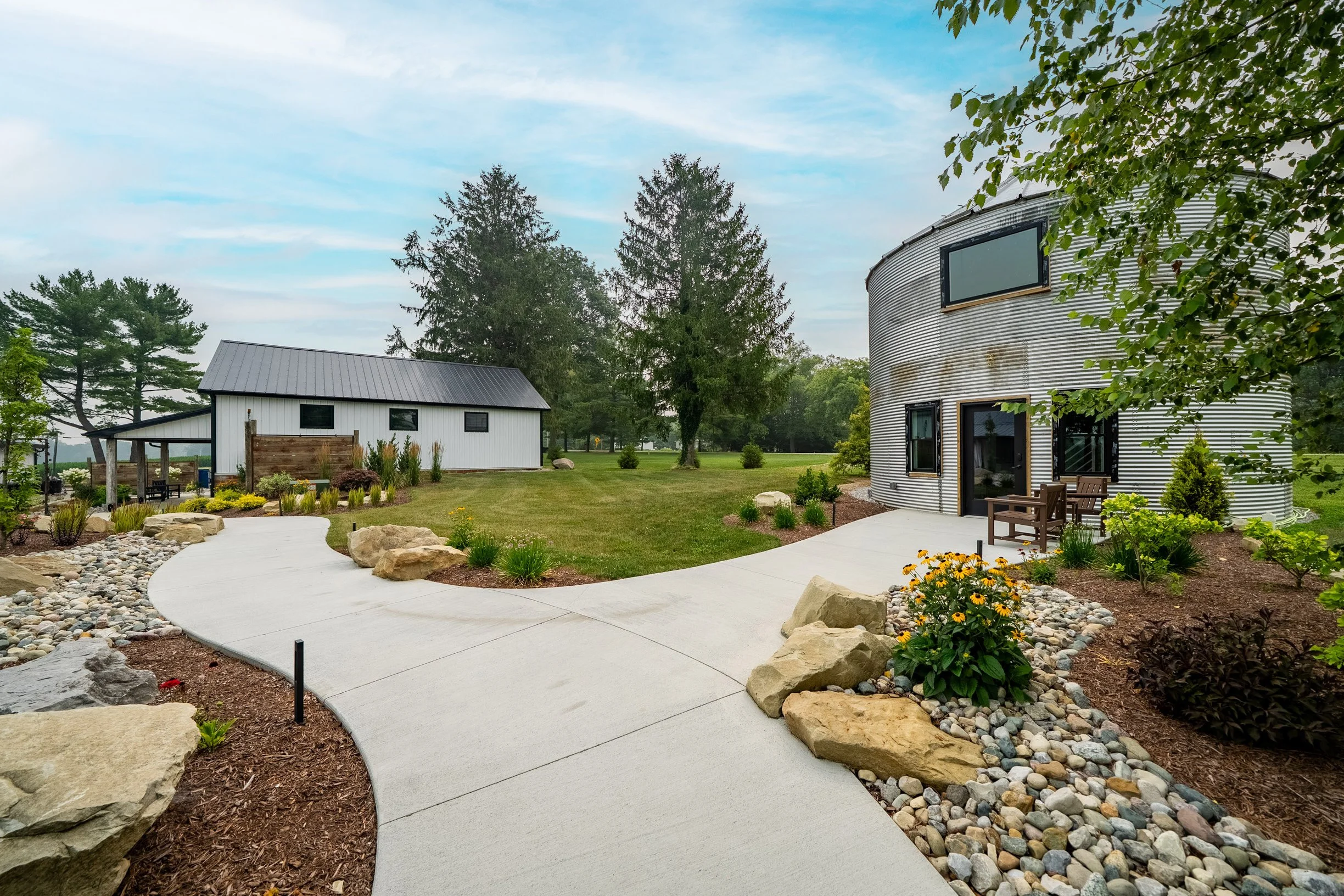 Garden with a concrete pathway, rocks, flowers, trees, a curved metal building, and a smaller white shed in the background under a cloudy sky.