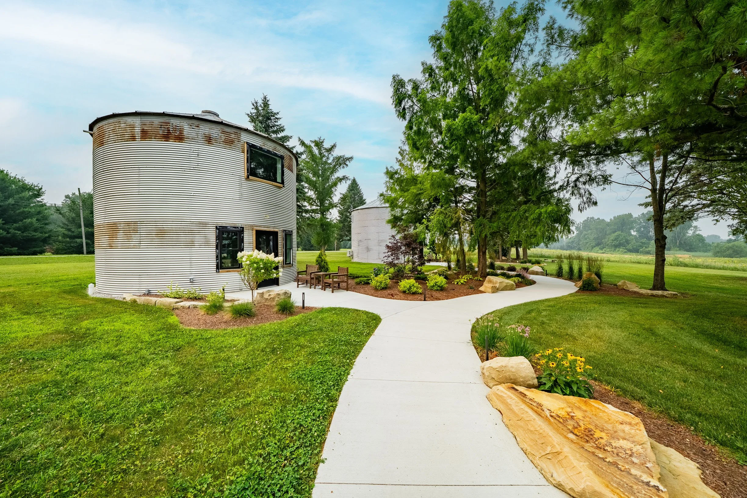 A backyard garden with a curved metal silo, a concrete pathway, green grass, trees, and outdoor seating.