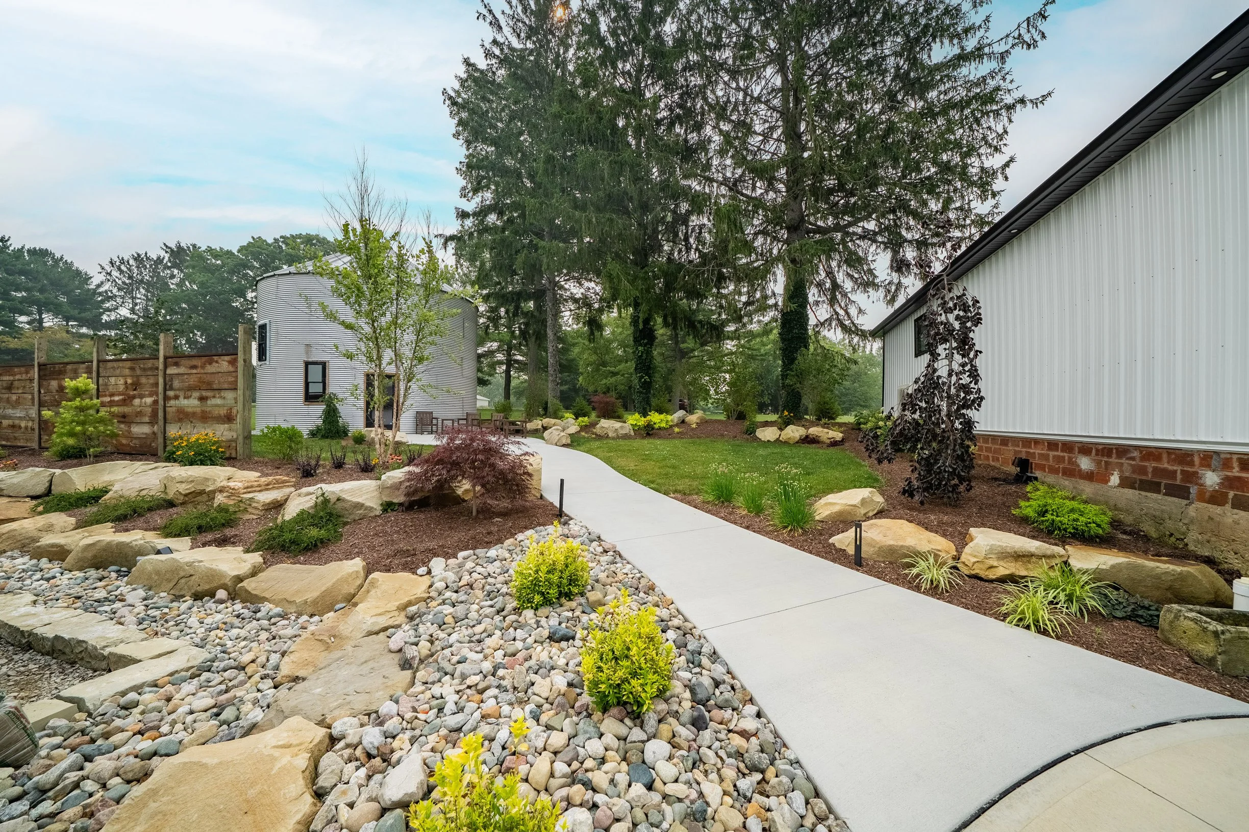 A landscaped backyard with a concrete pathway, rocks, small shrubs, and tall trees next to a metal building and a round silo.